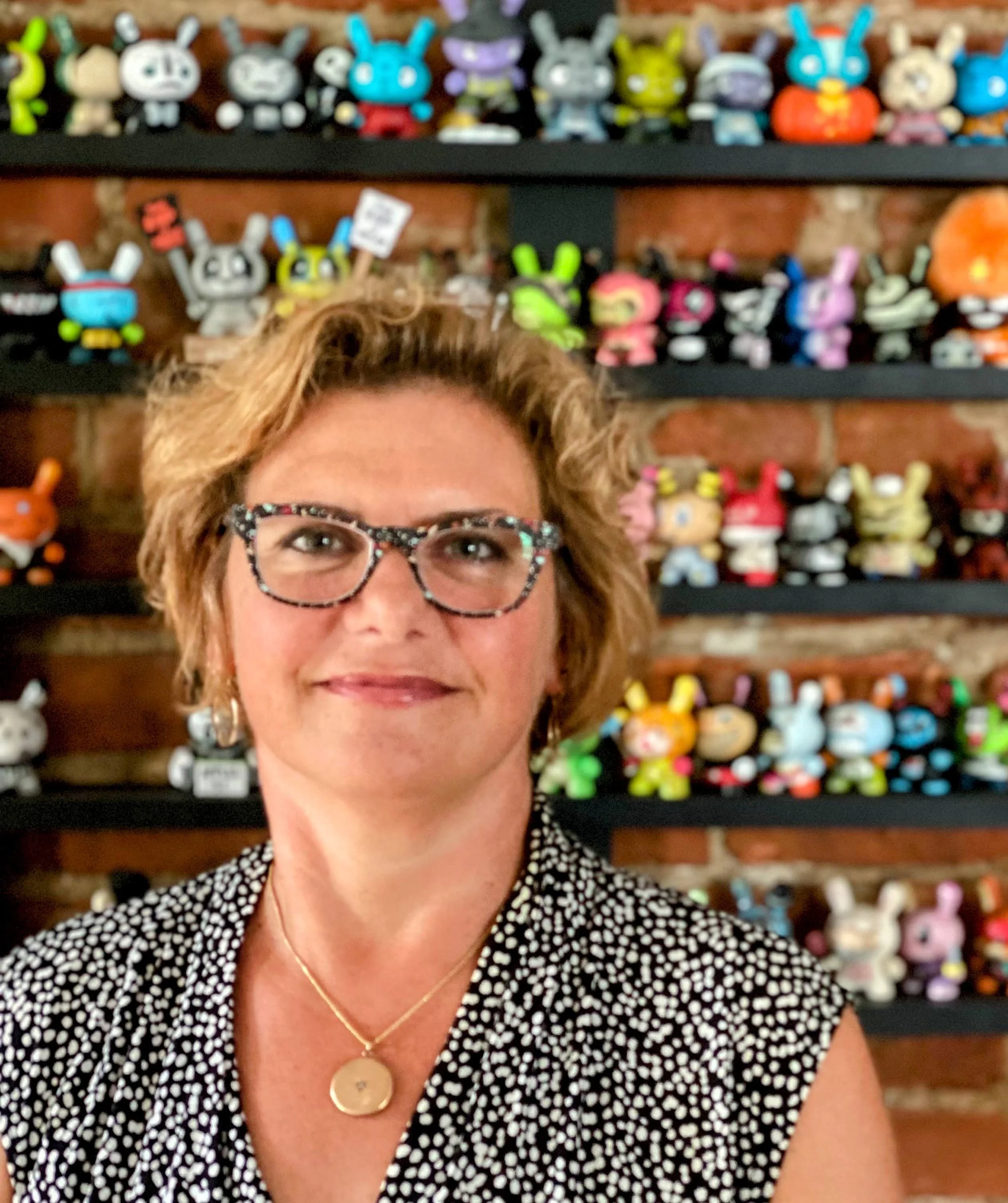 An image of a woman, wearing glasses and a polka dot shirt in front of a shelf of vinyl toys.