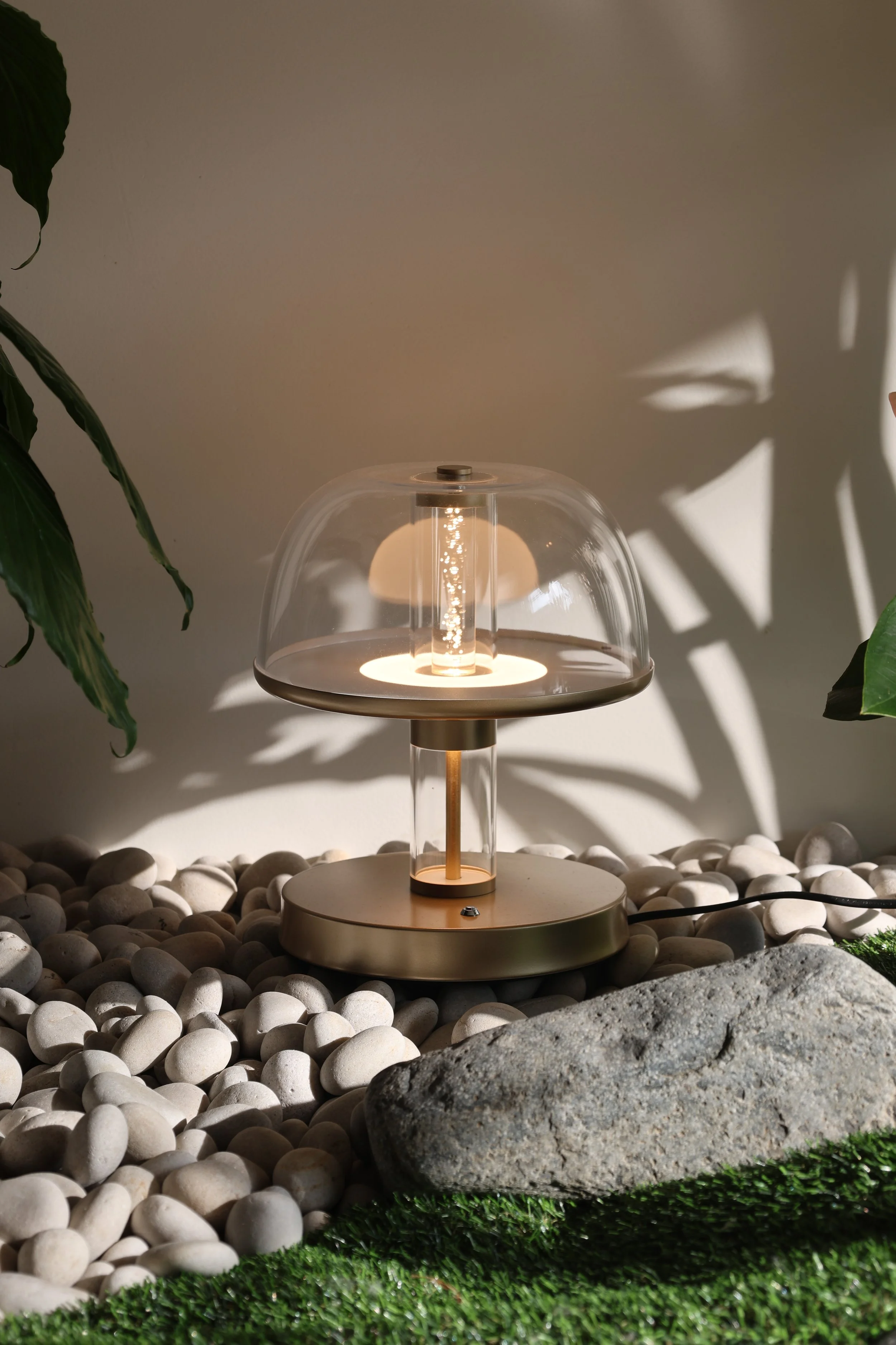 A modern table lamp with a transparent glass shade, sitting on a round beige base, surrounded by white pebbles, a large gray rock, green grass, and a wall with shadows from nearby plants.