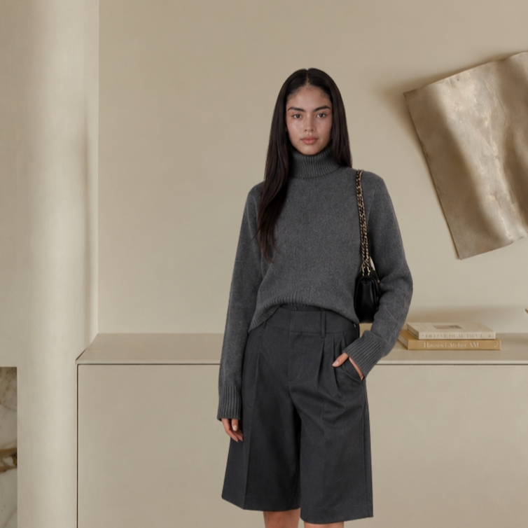 Woman wearing a gray turtleneck sweater and dark gray shorts, carrying a black shoulder bag with chain strap, standing in a neutral-toned room.