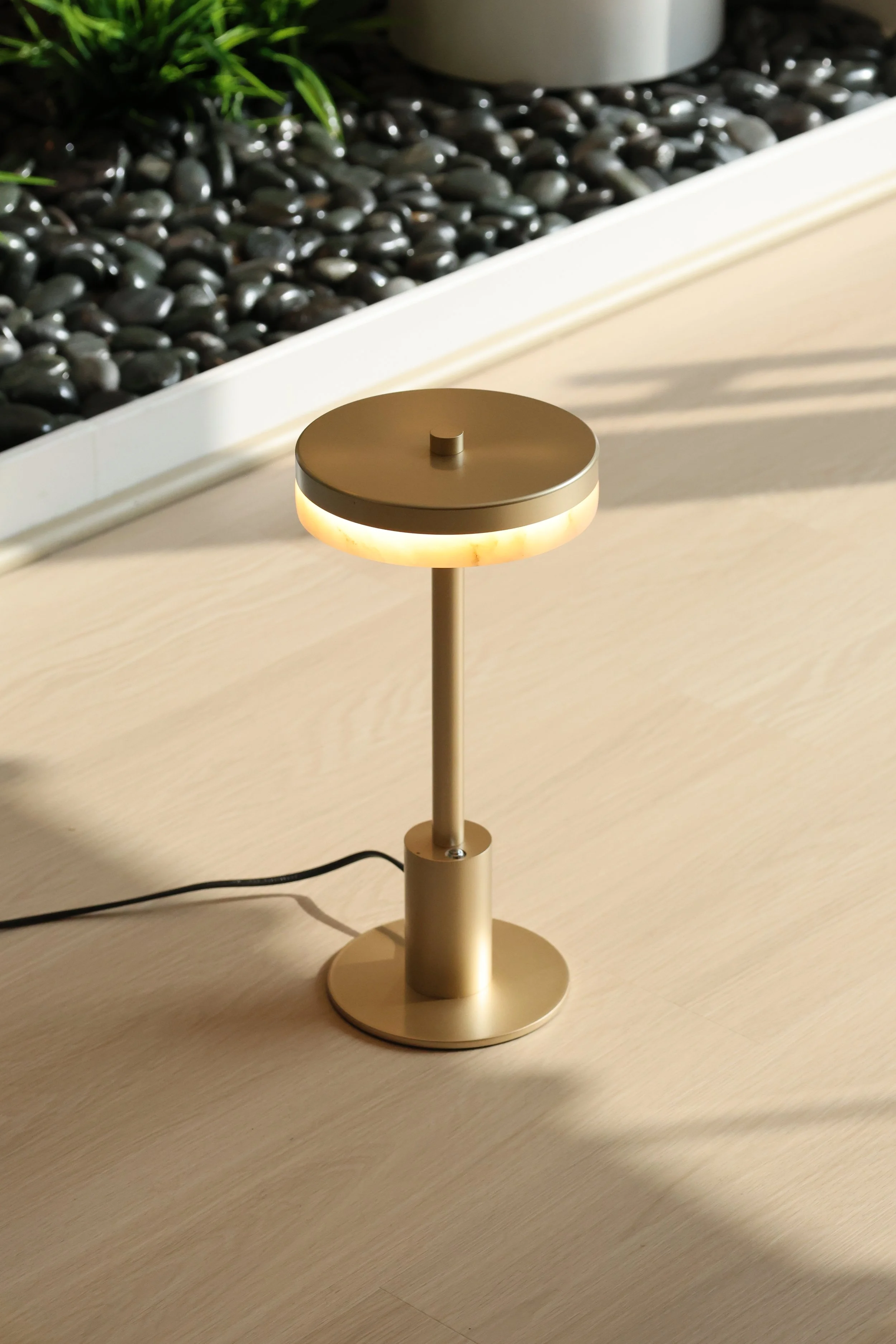 A modern table lamp with a round metallic top and glowing light underneath, placed on a wooden surface. In the background, there is a planter with green plants and black decorative stones.