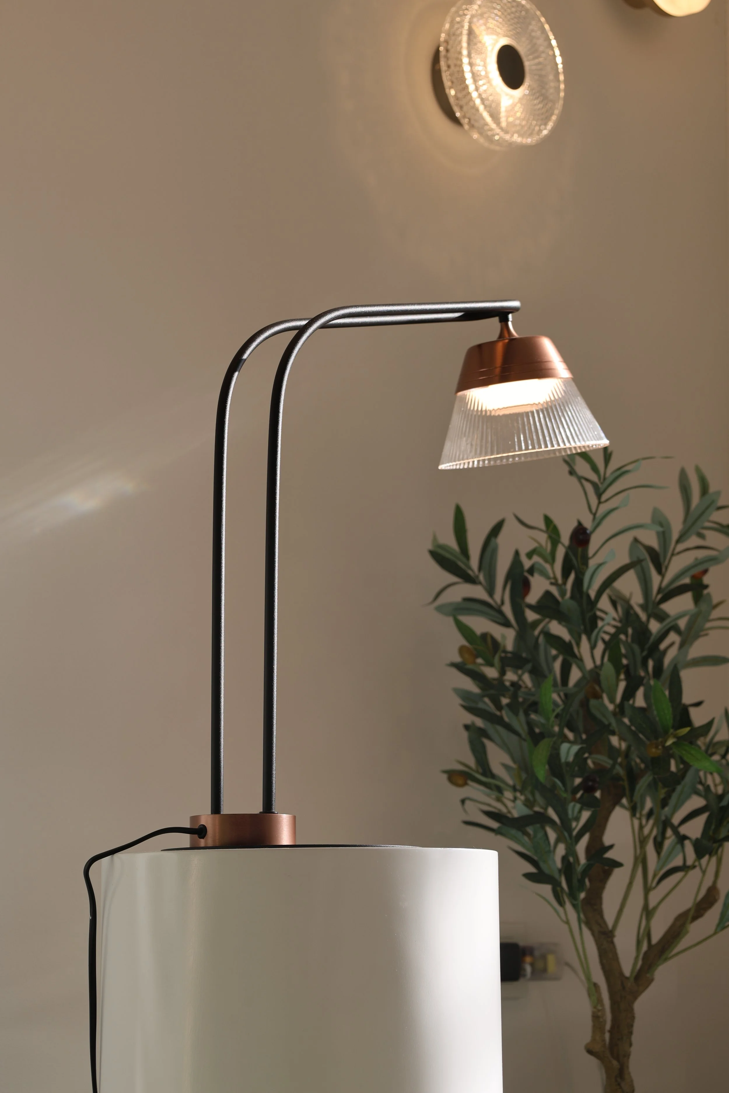 Modern table lamp with a metallic adjustable neck, a white cylindrical lampshade, and a decorative ceiling light fixture in the background. Green leafy plant near the lamp.