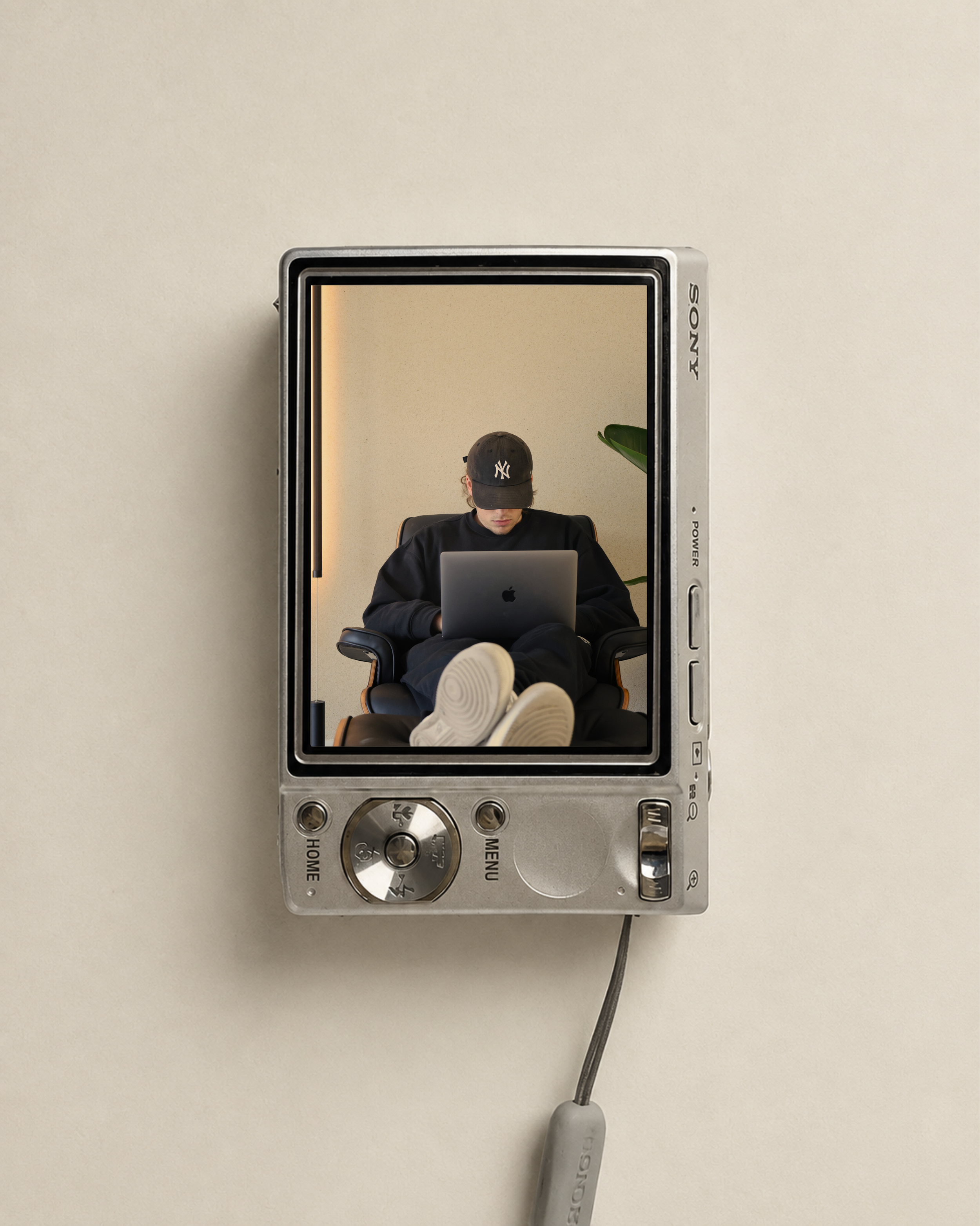 A person wearing a black hoodie and a black baseball cap is sitting in a black office chair with legs stretched out, working on a silver laptop. The person is inside a silver compact camera body that appears to be placed on a beige surface, with a visible black cord attached to the camera.