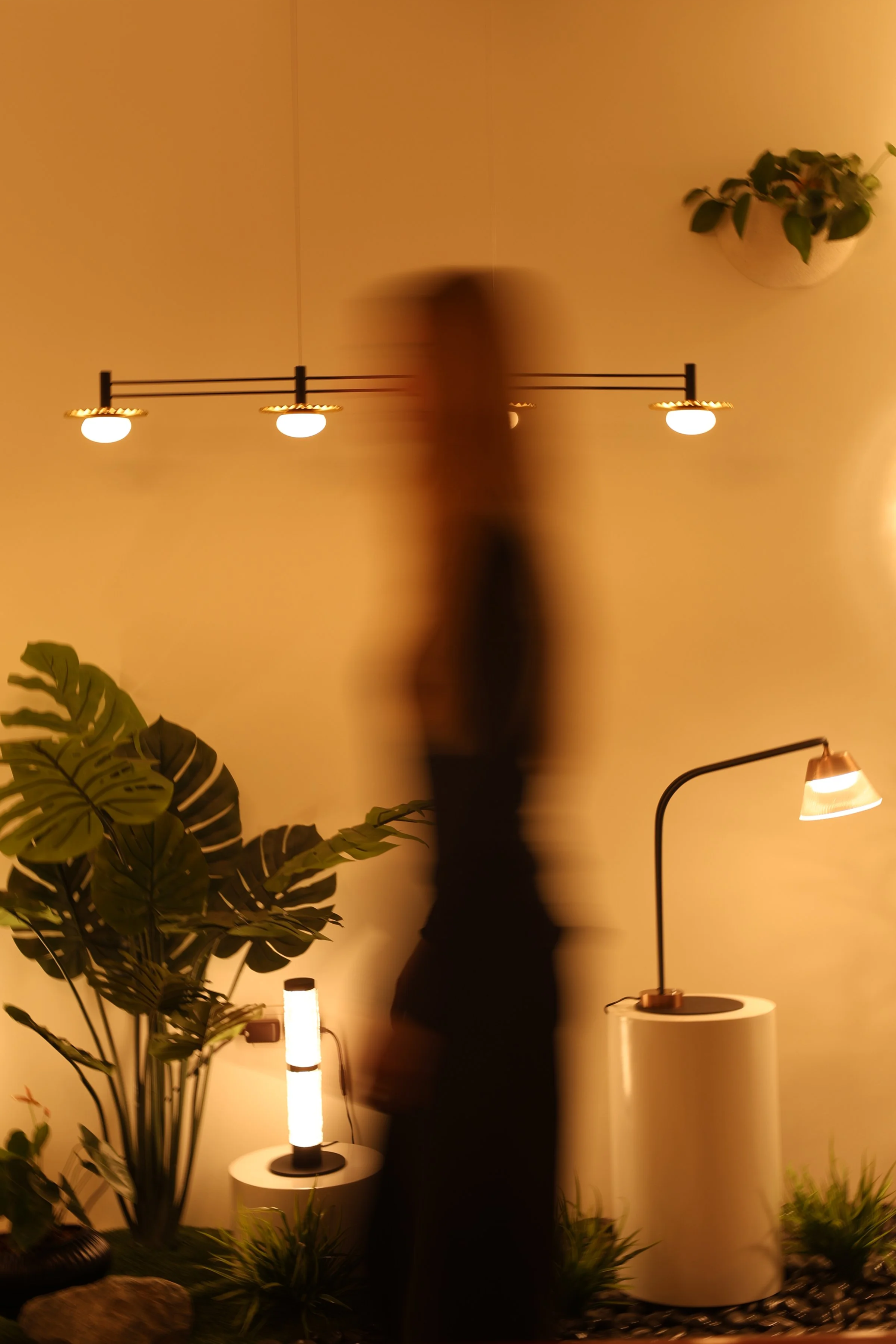 A blurred person in black standing in a warmly lit room with plants, a modern standing lamp, and ceiling lights.