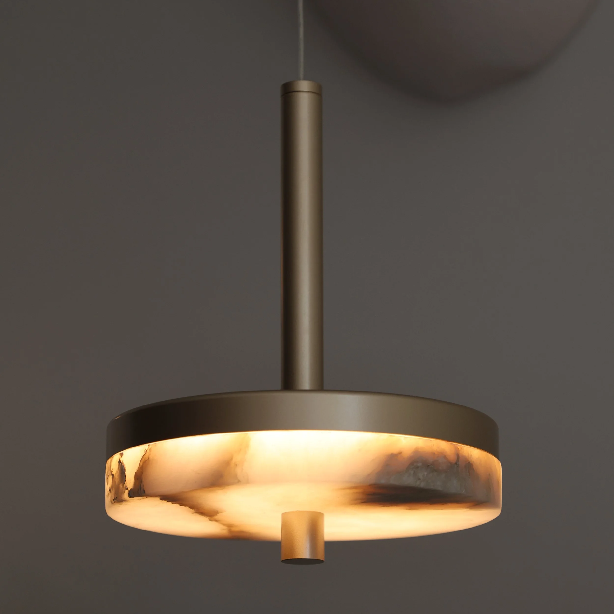 A modern pendant ceiling light with a cylindrical gold fixture emitting warm light, mounted against a gray ceiling.