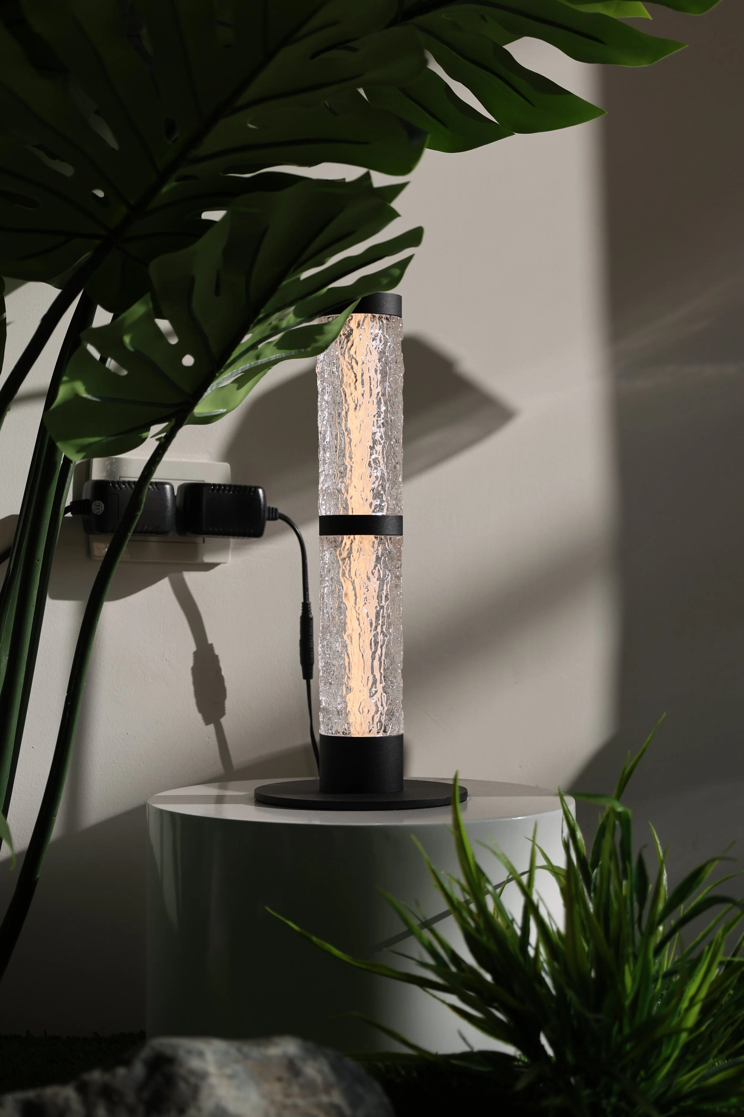 A modern table lamp with a textured, water-like glass body and a black base, situated on a small white table surrounded by green houseplants, with a power outlet and charger plugged in on the wall behind it.