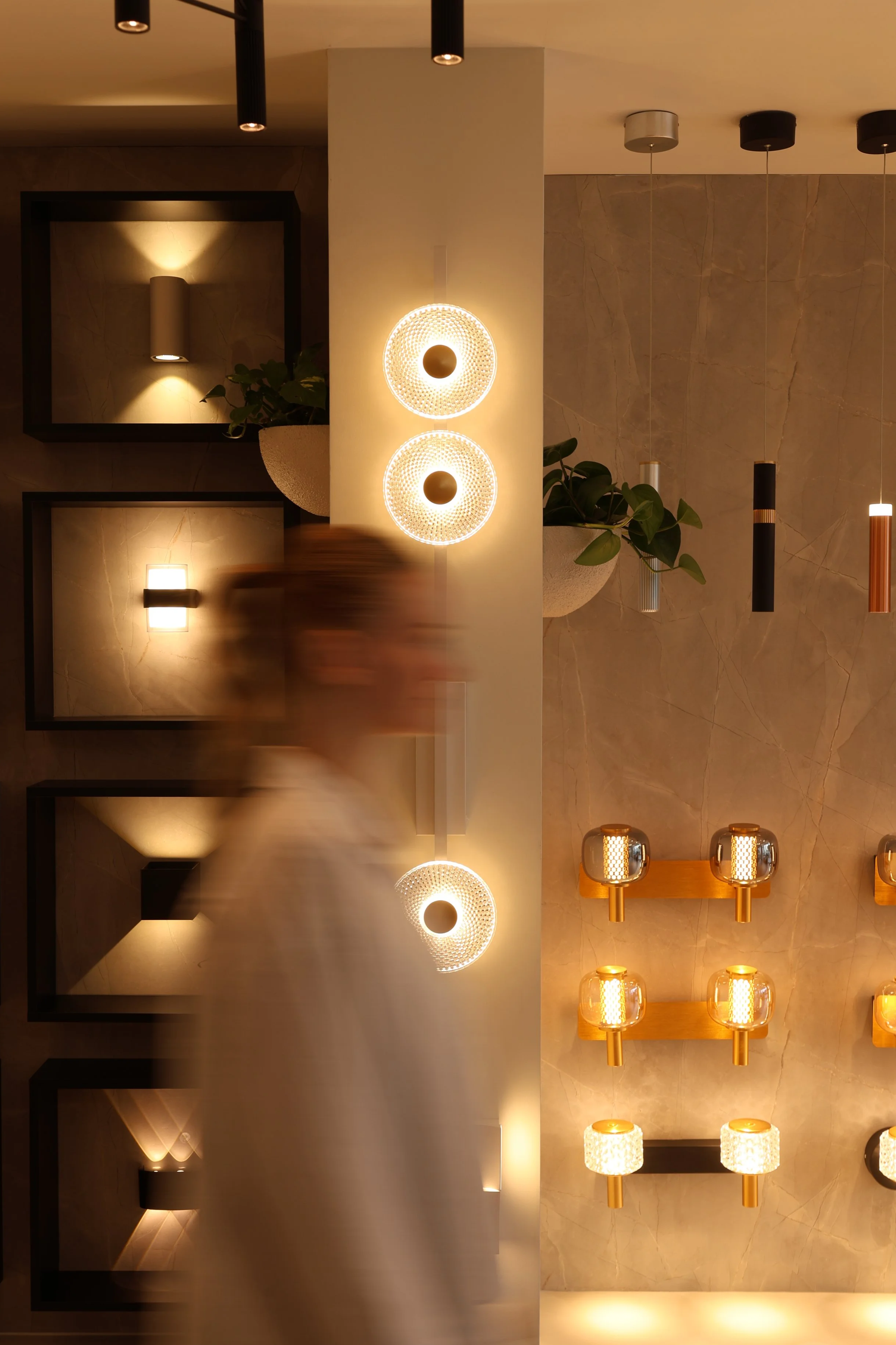 Various modern ceiling lights, including wall-mounted sconces, hanging pendant lamps, and decorative fixtures, on a beige marble wall with potted plants.