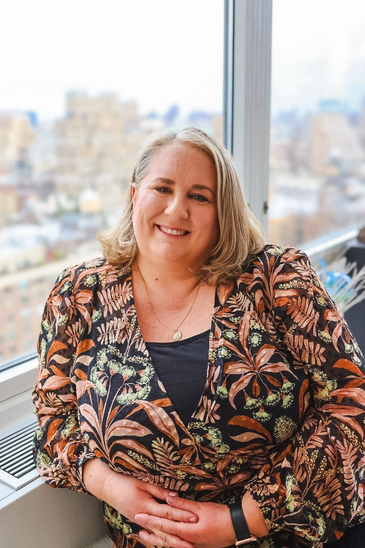 Woman smiling in floral shirt with window and cityscape in background.
