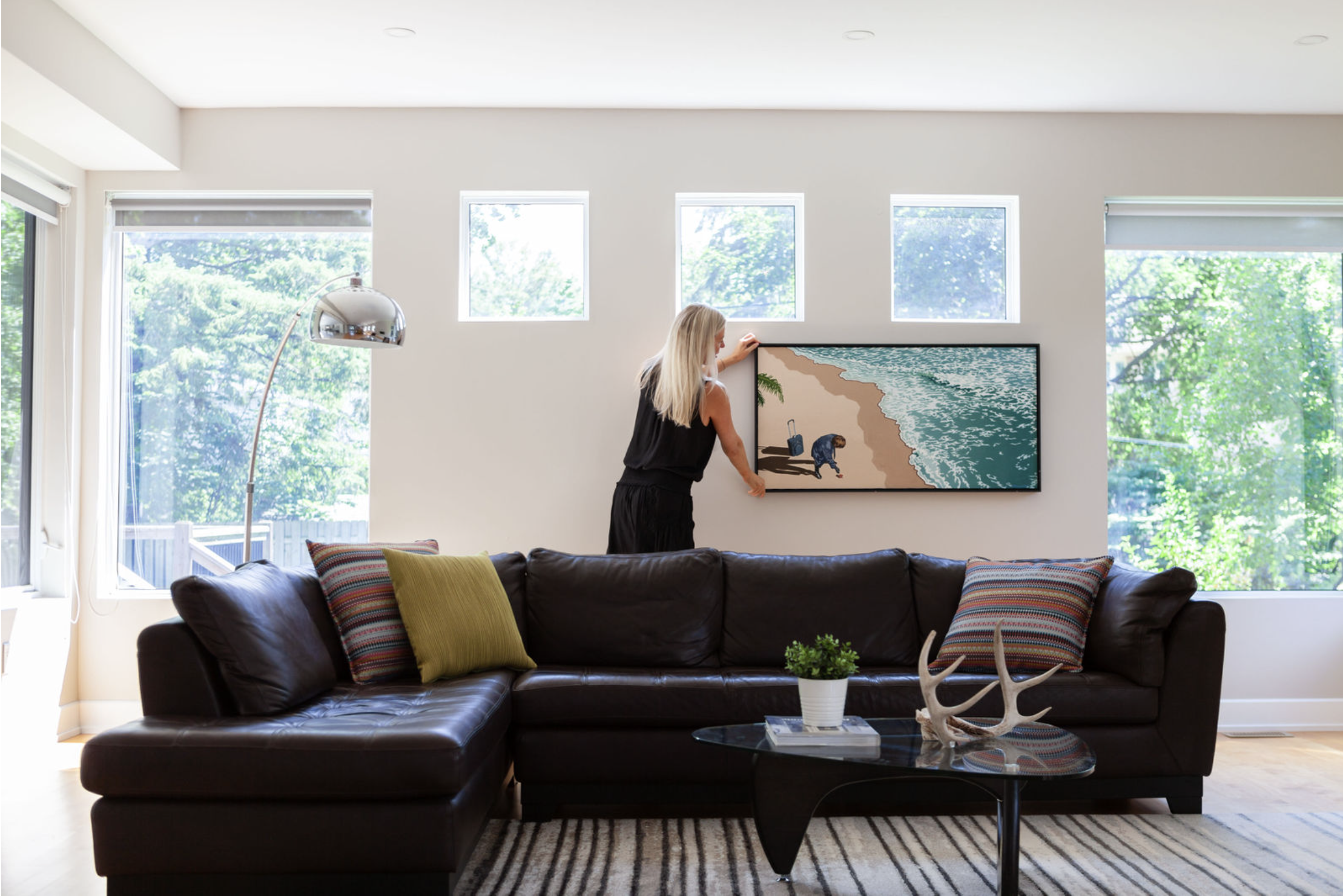 Woman hanging a framed beach scene painting on a white living room wall with a large brown leather sofa and decorative pillows, a black glass coffee table with a plant and antler decor, and large windows with natural sunlight.