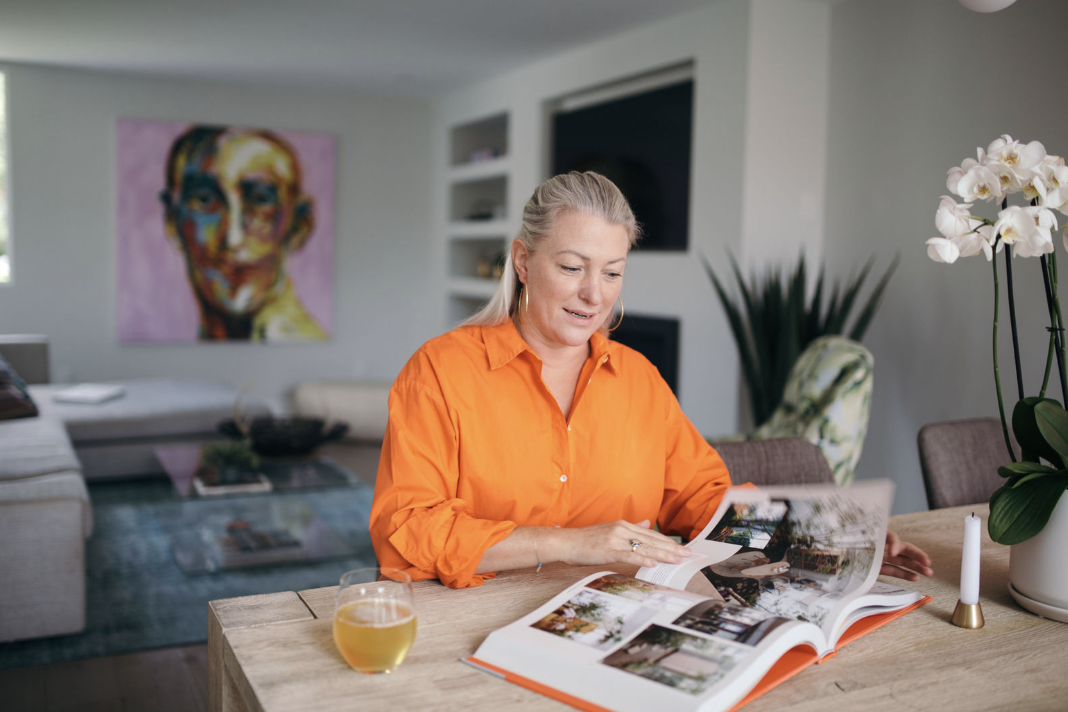 Woman in an orange shirt reading a magazine at a wooden table, with a glass of drink, a candle, and a potted plant nearby, in a modern living room.