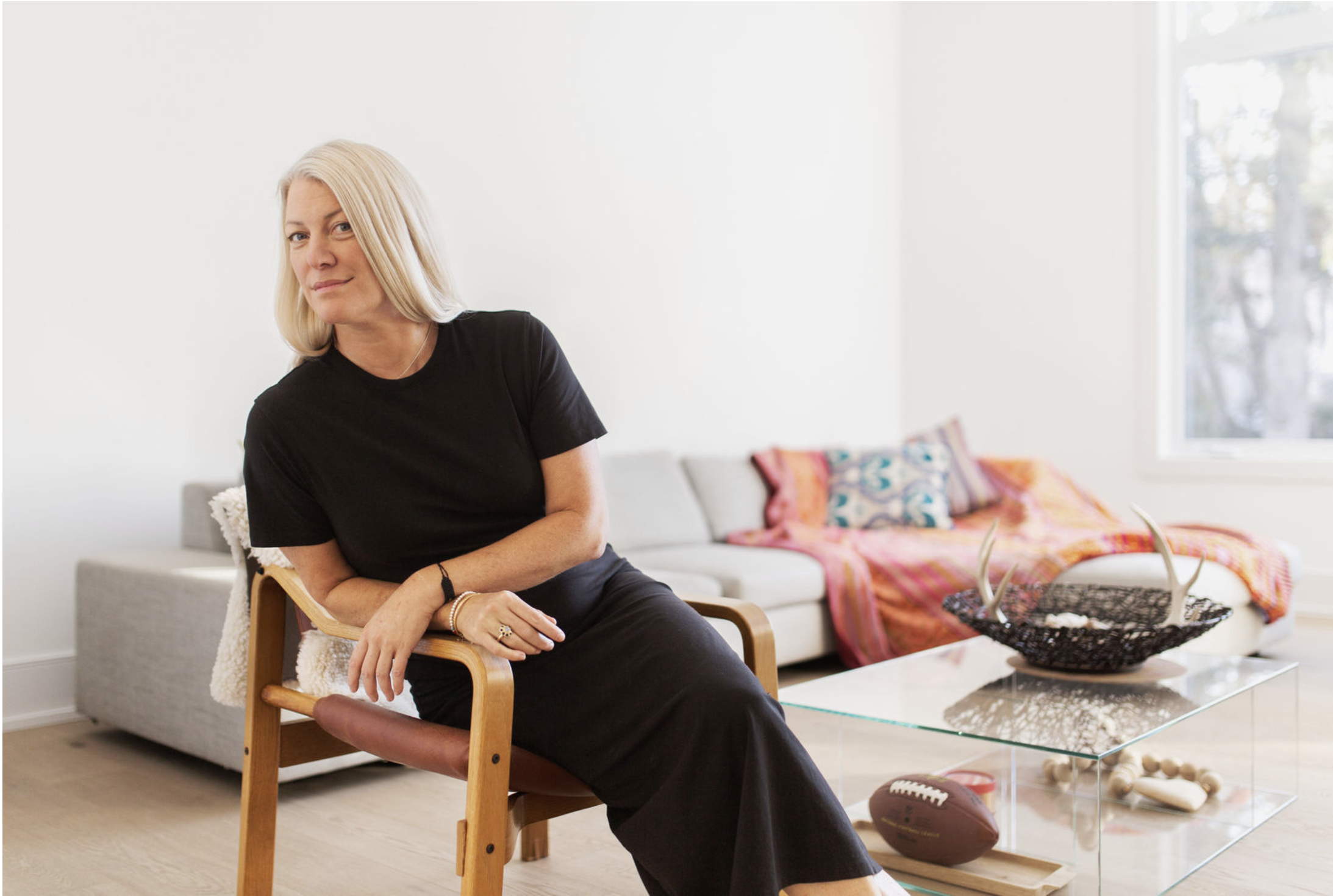 A woman with blonde hair in a black t-shirt and black pants sitting on a wooden and leather chair in a bright living room. A white sofa with colorful pillows and blanket is in the background, with a glass coffee table holding a decorative bowl and footballs. A large window lets in natural light.
