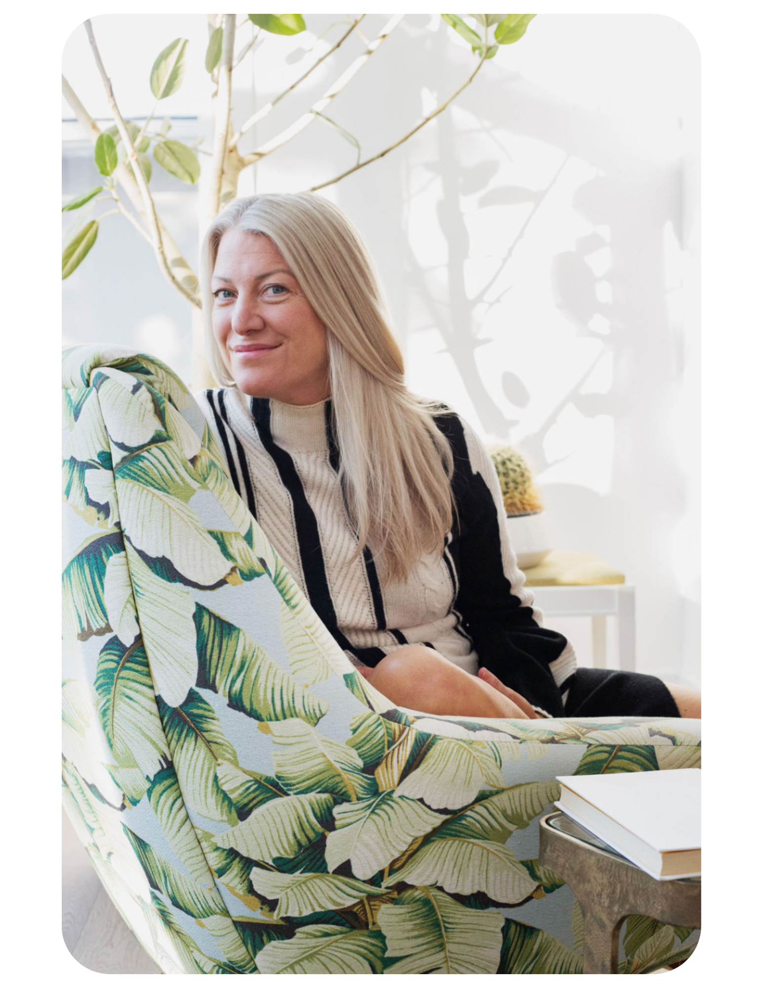 A woman with blonde hair sitting on a chair with tropical leaf print fabric, smiling in a bright room with natural light and houseplants.