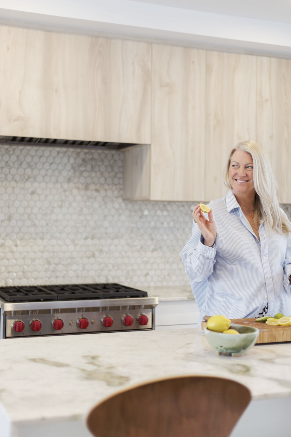 A woman with blonde hair smiling and holding a lemon wedge in a modern kitchen with light wood cabinets, a white marble countertop, and a bowl of lemons.