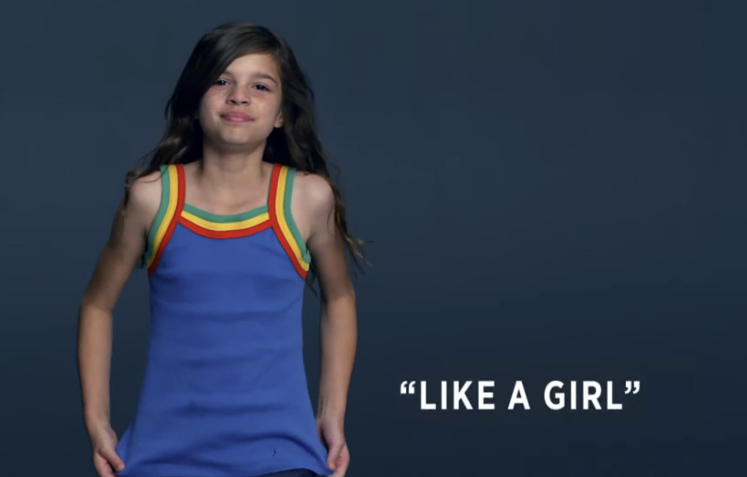 Young girl with long dark hair wearing a colorful striped dress, standing against a dark background with the caption 'LIKE A GIRL'.