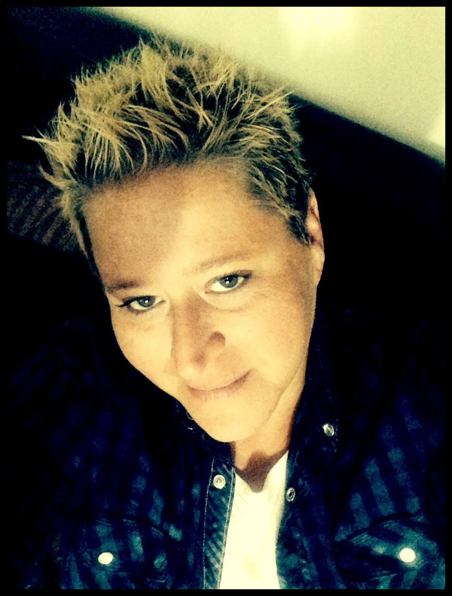 A young person with spiky blond hair, blue eyes, and fair skin, wearing a dark plaid shirt and a white T-shirt, lying down and looking at the camera.