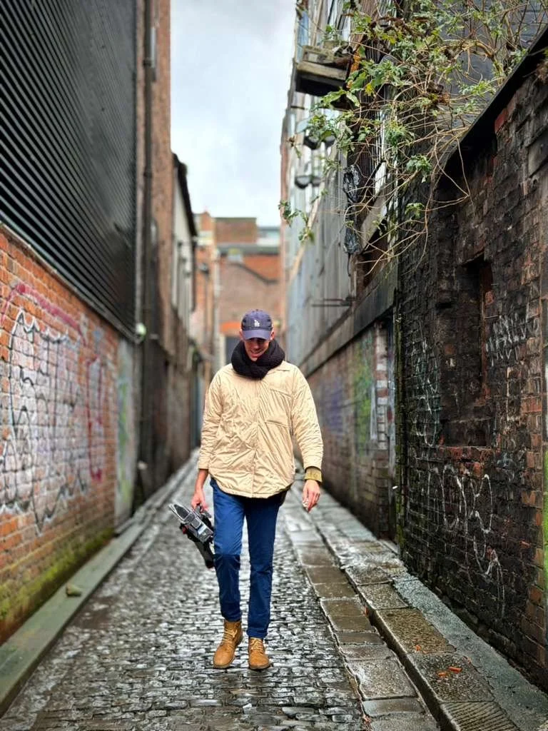 A person walking down a narrow, wet alleyway with brick walls on either side, holding a camera in their right hand.