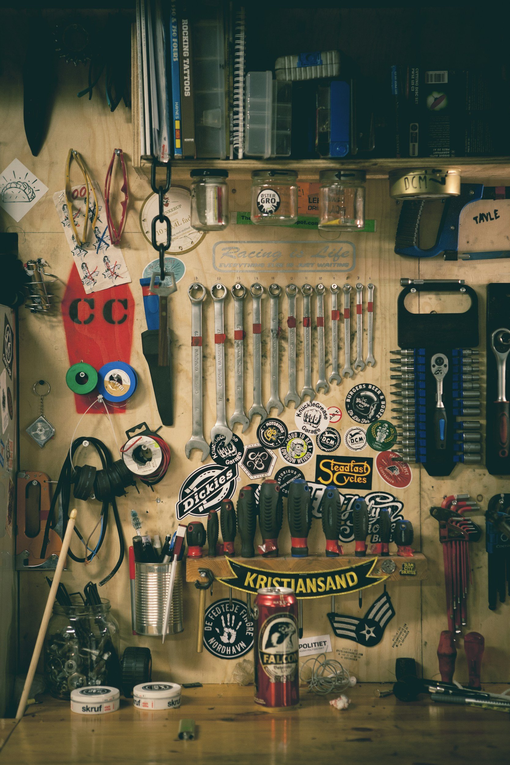 A well-organized workbench with tools, stickers, and small parts, including screwdrivers, wrenches, and scissors, with a can of Falcon beer on the surface.