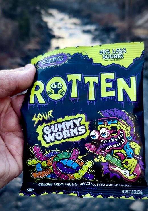 A person holding a bag of Rotten Sour Gummy Worms candy with colorful cartoon worms and a monster illustration on the packaging.