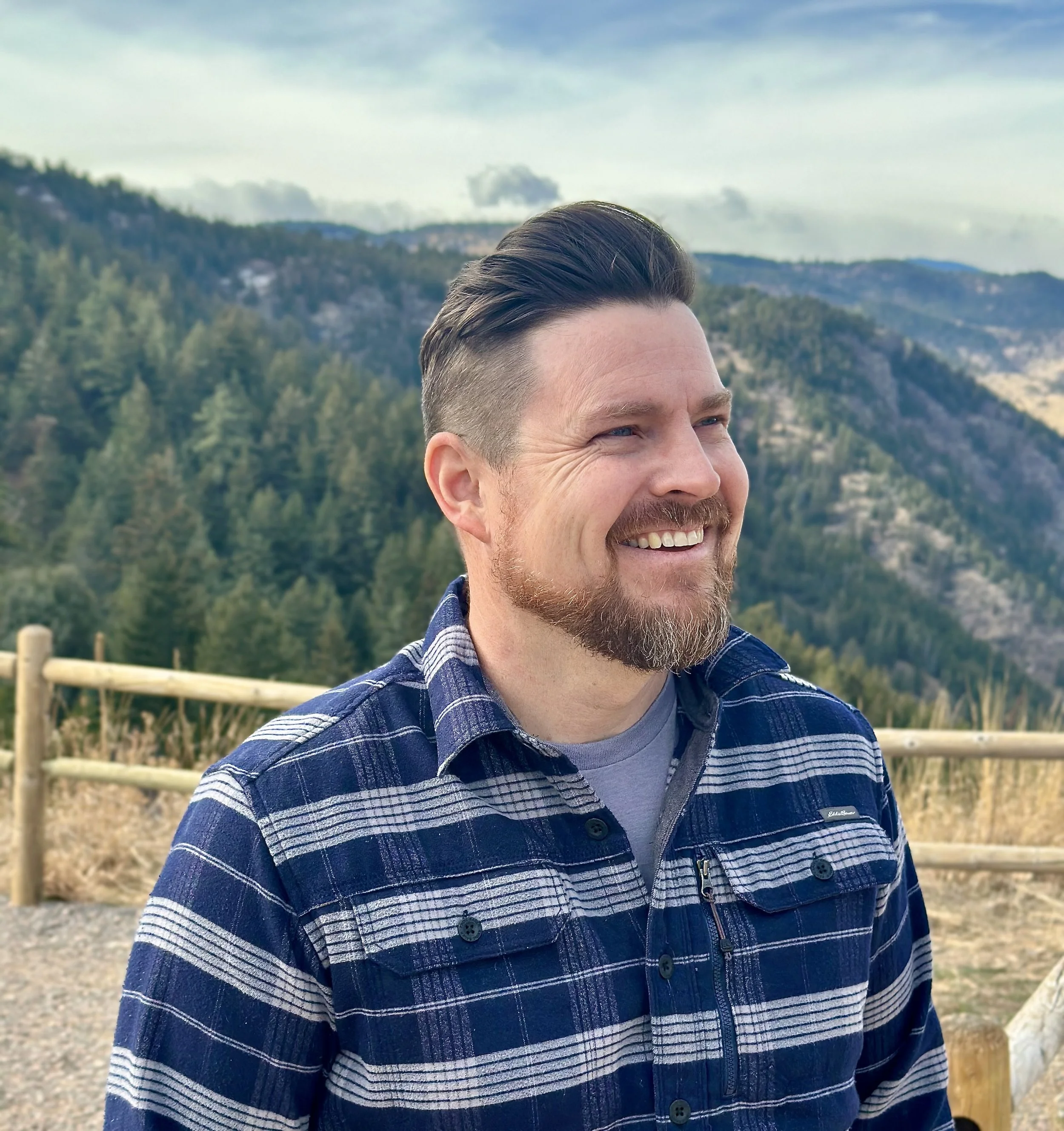 A smiling man with dark hair and a beard standing outside with a landscape of mountains and trees in the background.