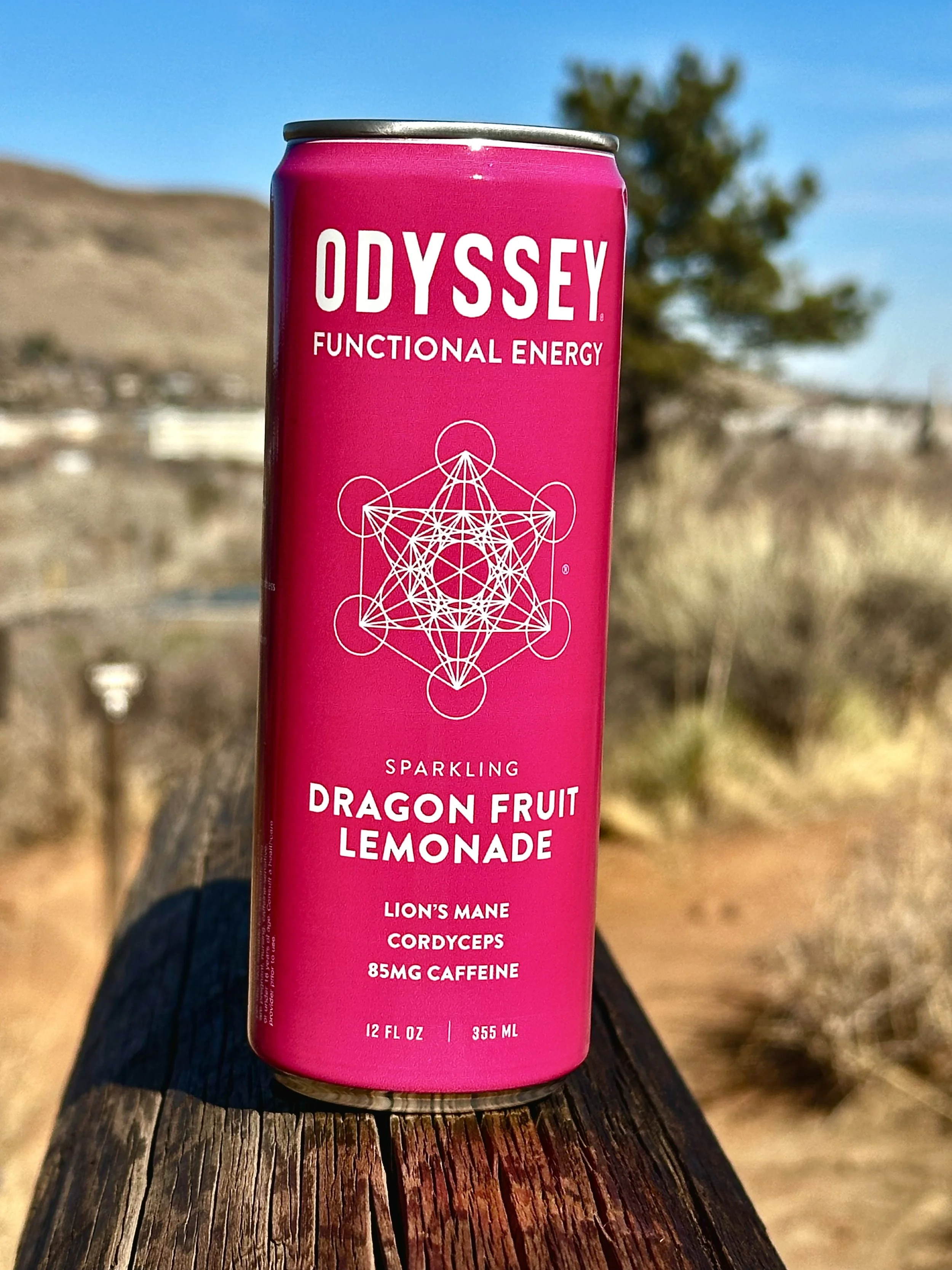 A pink can of Odyssey functional energy sparkling dragon fruit lemonade with geometric artwork on the front, placed outdoors on a wooden surface with a natural background and clear sky.
