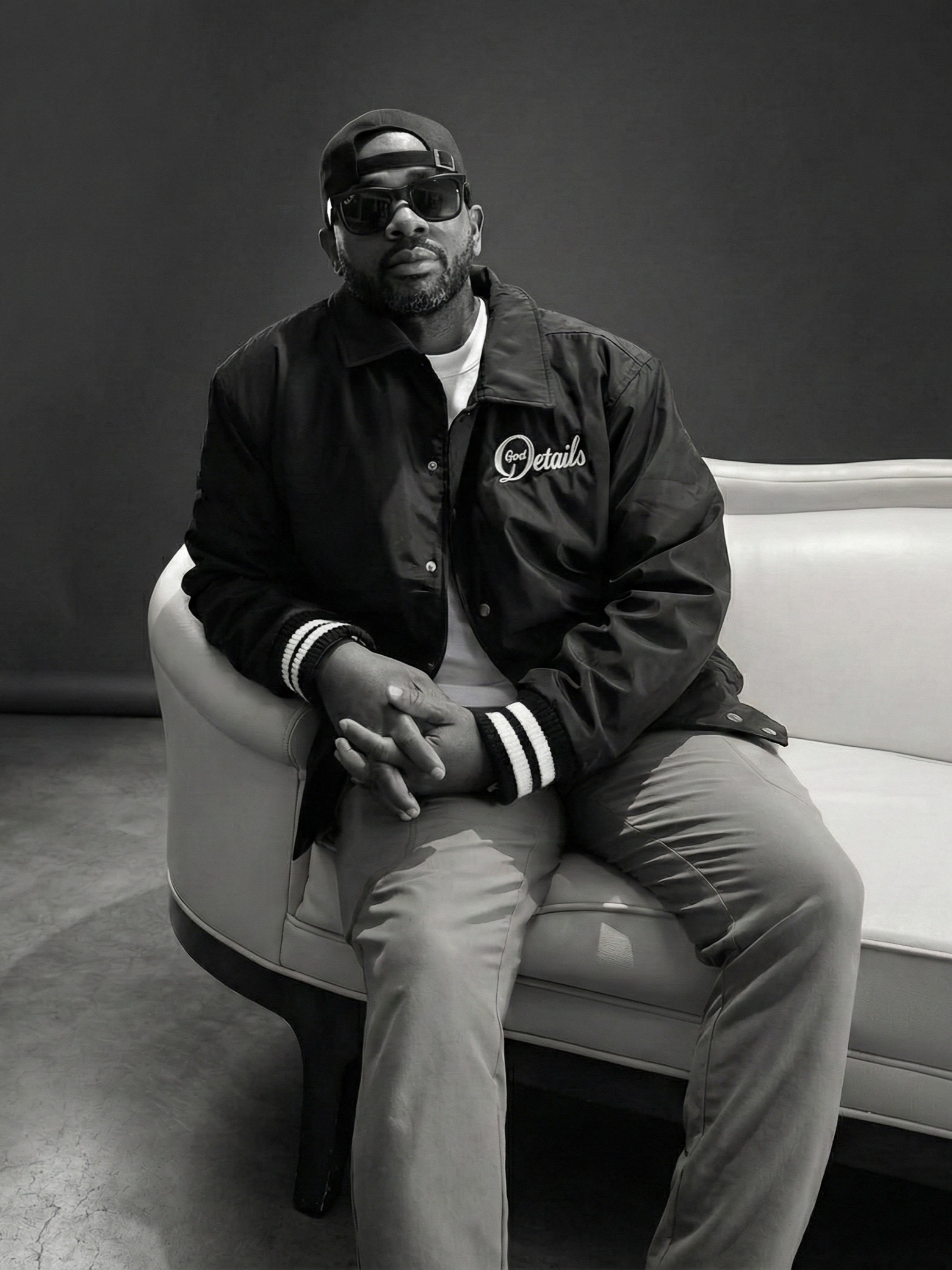 A man with a beard wearing sunglasses, a backward cap, a black jacket with 'God Details' embroidered, sitting on a white sofa with arms crossed, in a plain indoor setting.