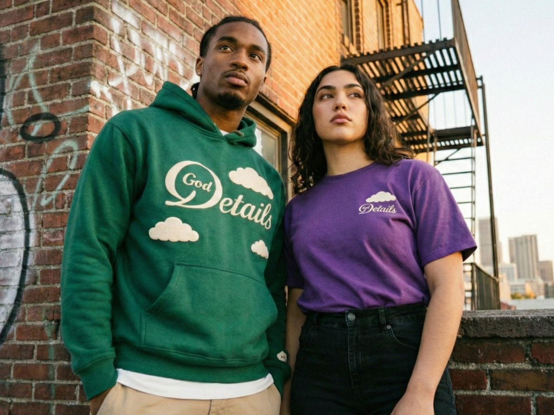 Two young adults standing outdoors on a city rooftop with city skyline in background. The man is wearing a green hoodie with the words "God Details" and clouds printed on it. The woman is wearing a purple T-shirt with smaller clouds and the word "Det