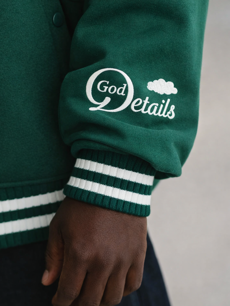 Close-up of a person wearing a green jacket with white text and a cloud logo that reads 'God Details'. The jacket has a striped green and white cuff.