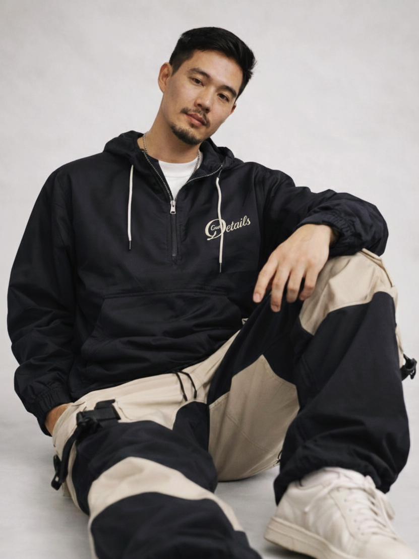 A young man with dark hair and a beard sits on the floor against a plain background, wearing a black hoodie with white drawstrings and beige and black cargo pants, gazing at the camera.