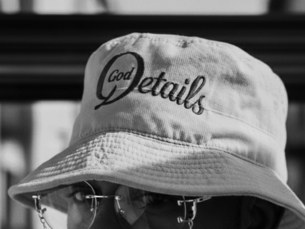 Person wearing a bucket hat with the embroidered text "God Details," wearing round glasses, with a background of horizontal black slats.