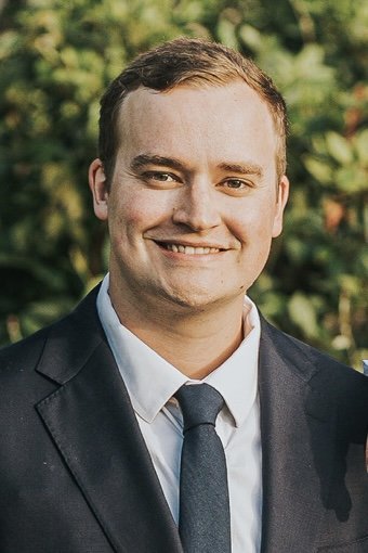 Portrait of a young man in a suit smiling outdoors with greenery in the background.