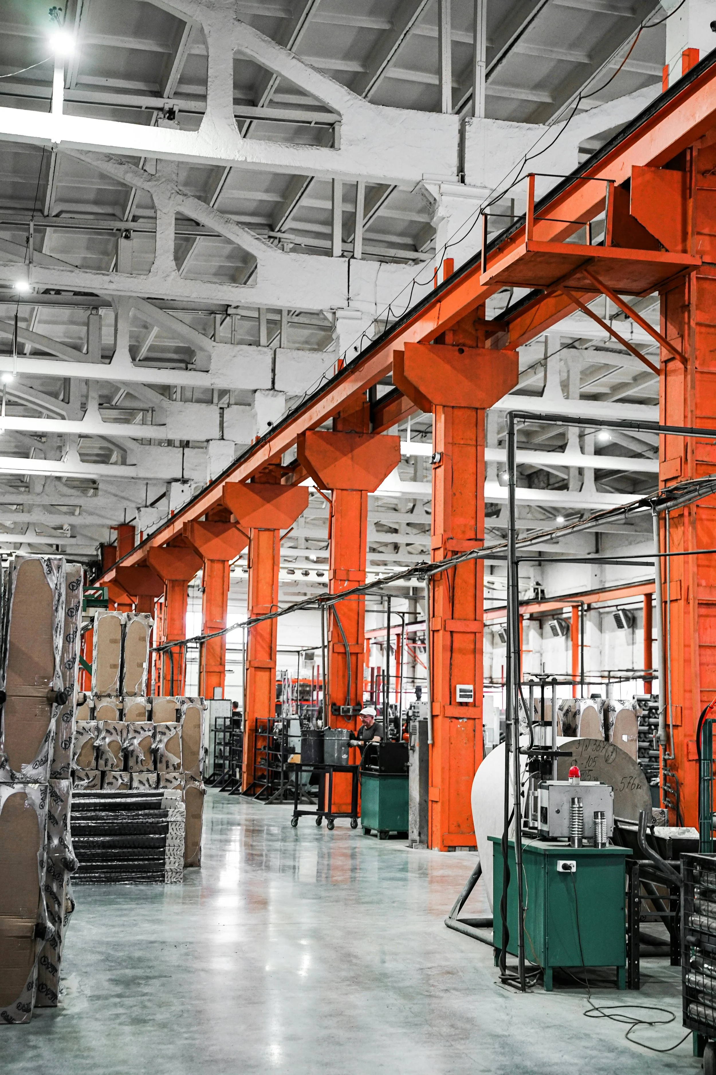 Interior of a factory or warehouse with orange overhead cranes, some equipment, and a worker wearing a hard hat.