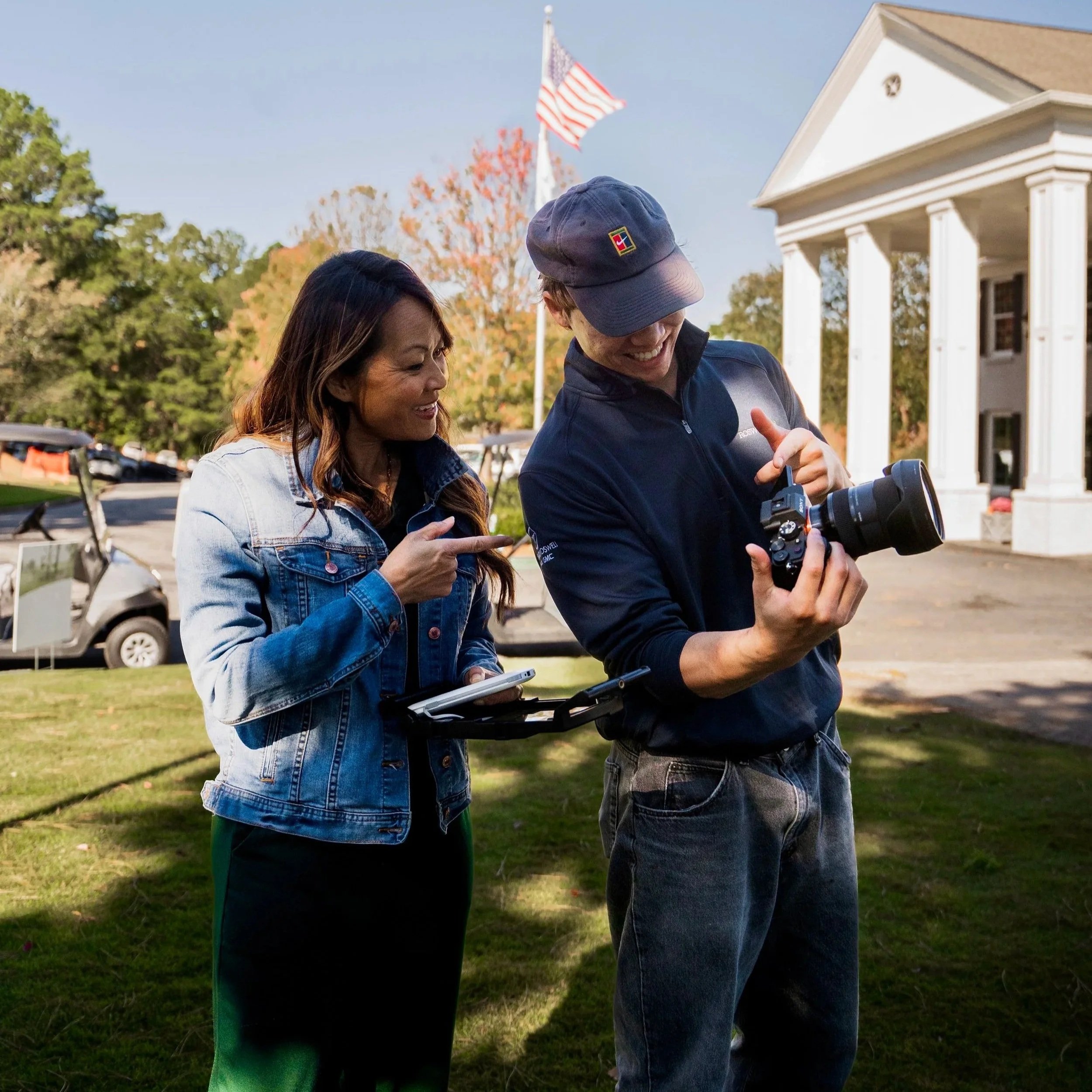 A woman in a denim jacket and a man in a dark jacket and baseball cap are outside, smiling and looking at a camera the man is holding. There is a white building with columns and a flagpole with an American flag in the background, along with trees and parked golf carts.