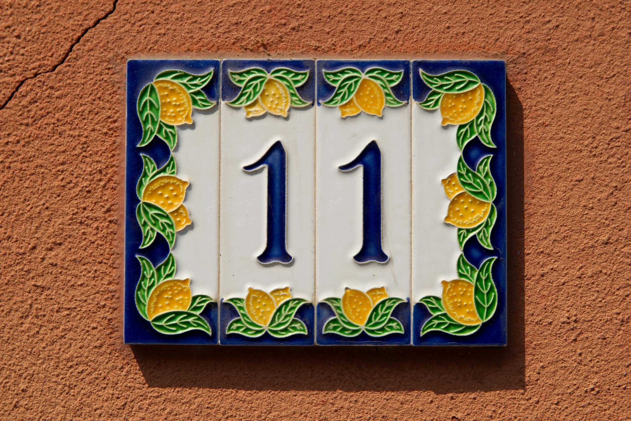 Decorative house number plaque with the number 111, featuring a border of yellow lemons and green leaves on a white background, mounted on a burnt orange wall.
