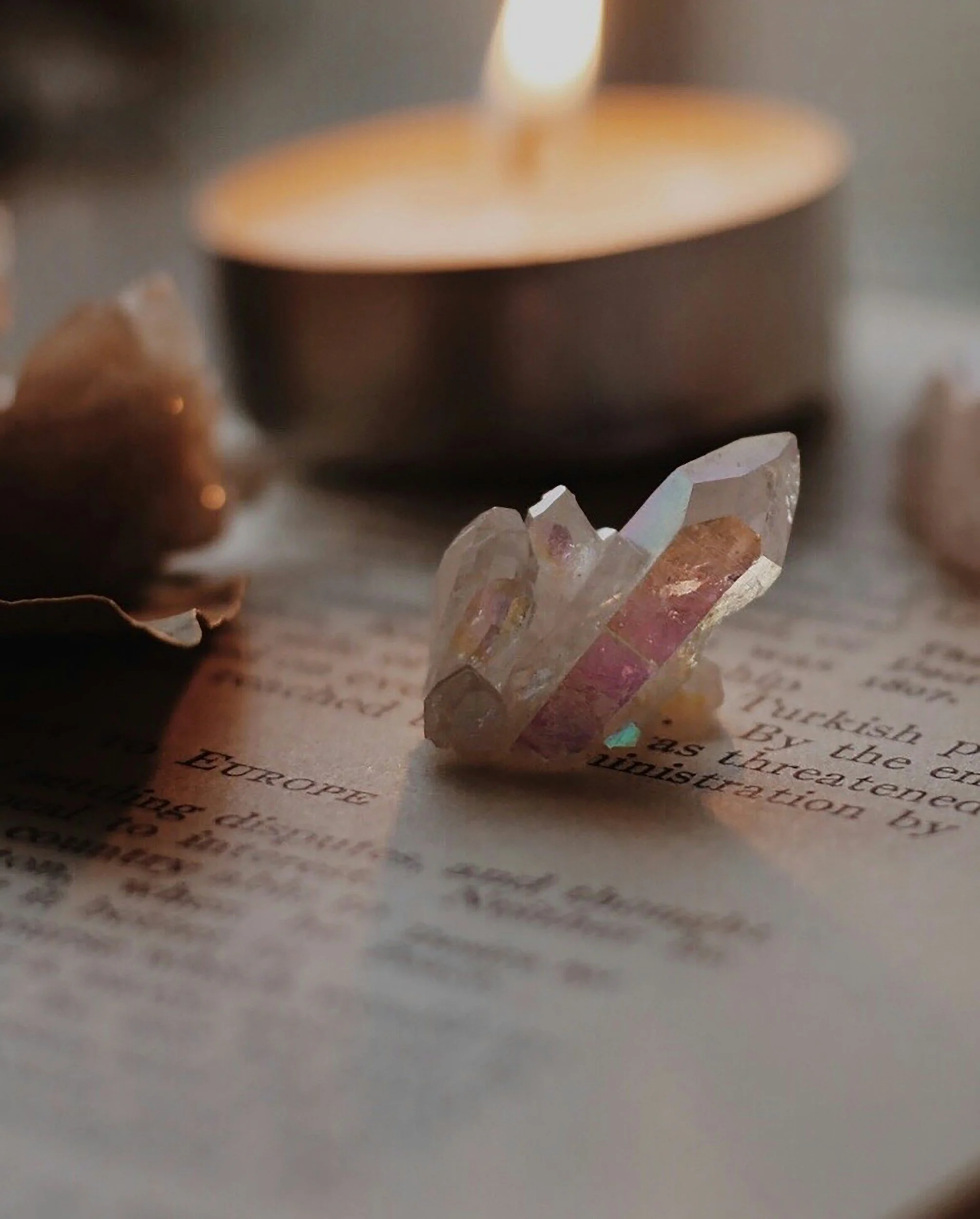 A small, clear quartz pendant with pink and purple hues resting on an open book with typed text, alongside dried flower petals and a lit candle in the background.