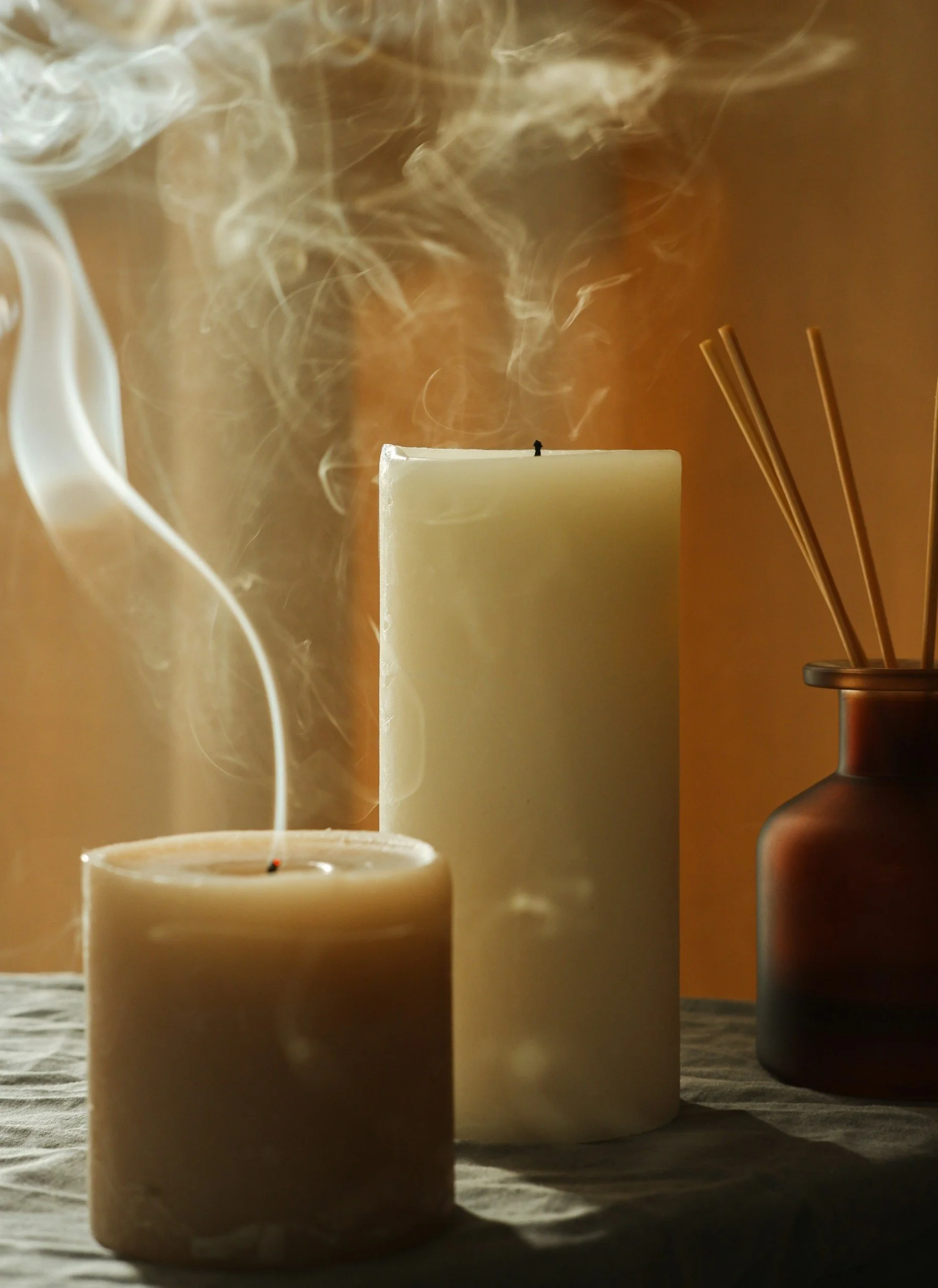 Two burning candles, one short and wide, and one tall and cylindrical, with wisps of smoke rising from the wicks. There is a brown vase with reed diffusers on the right, on a textured surface with an orange background.