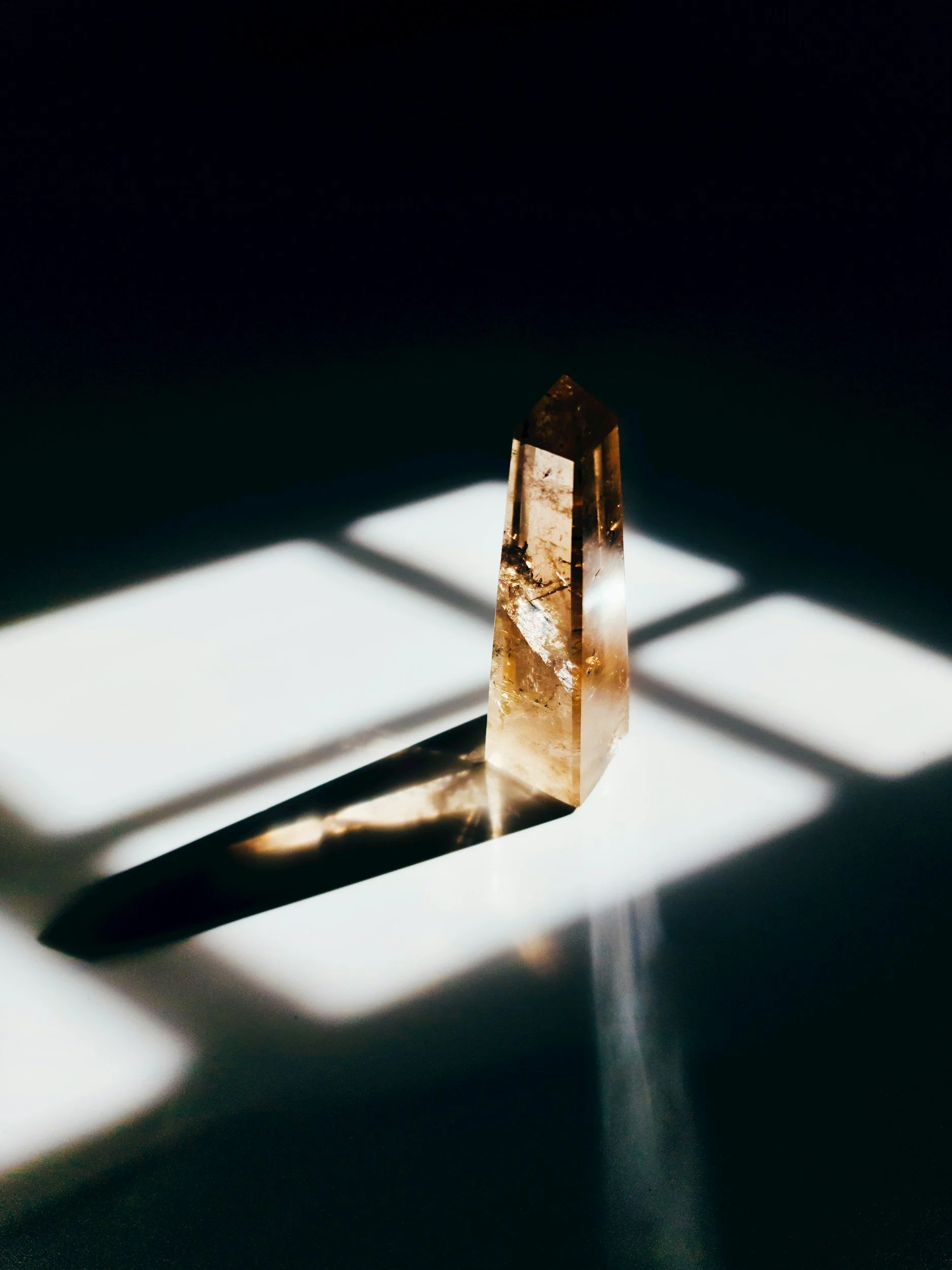 A clear, rectangular crystal standing upright on a white surface, with shadows and light creating patterns around it.
