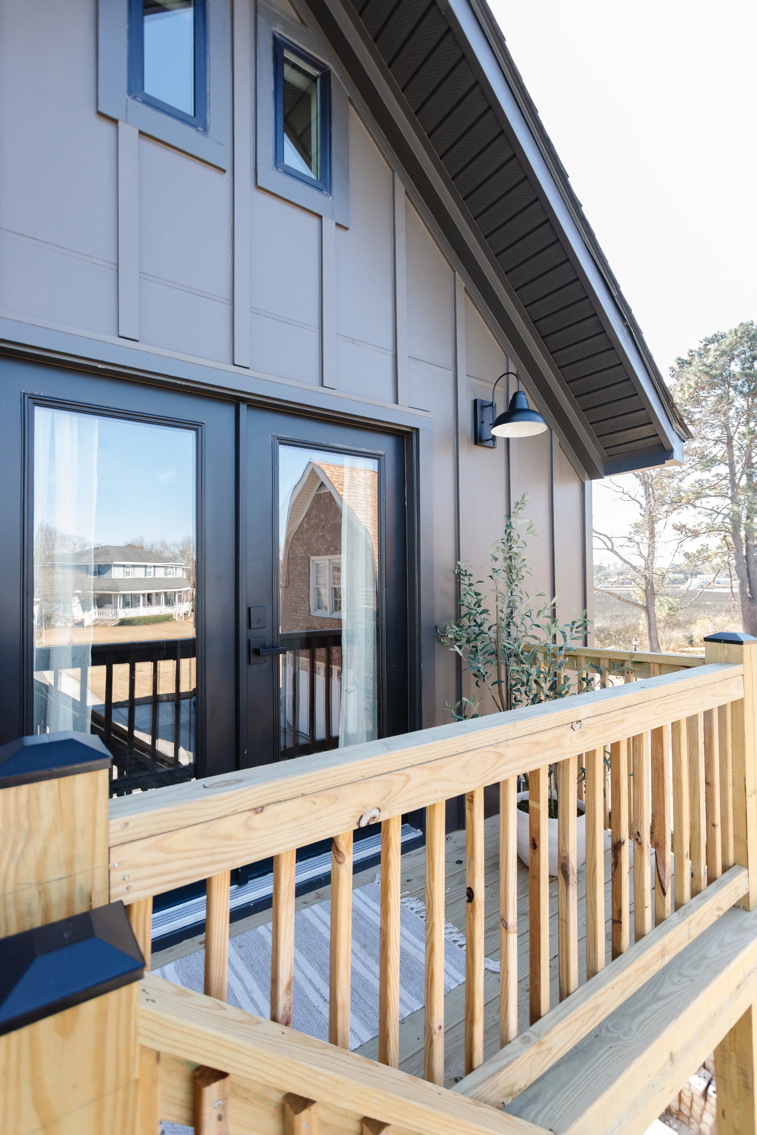 A wooden deck with railing, attached to a house with gray siding and small black-framed windows. There is a door with glass panes and a curtain, and a black outdoor wall-mounted light fixture. Outside, there is a view of neighboring houses and trees.