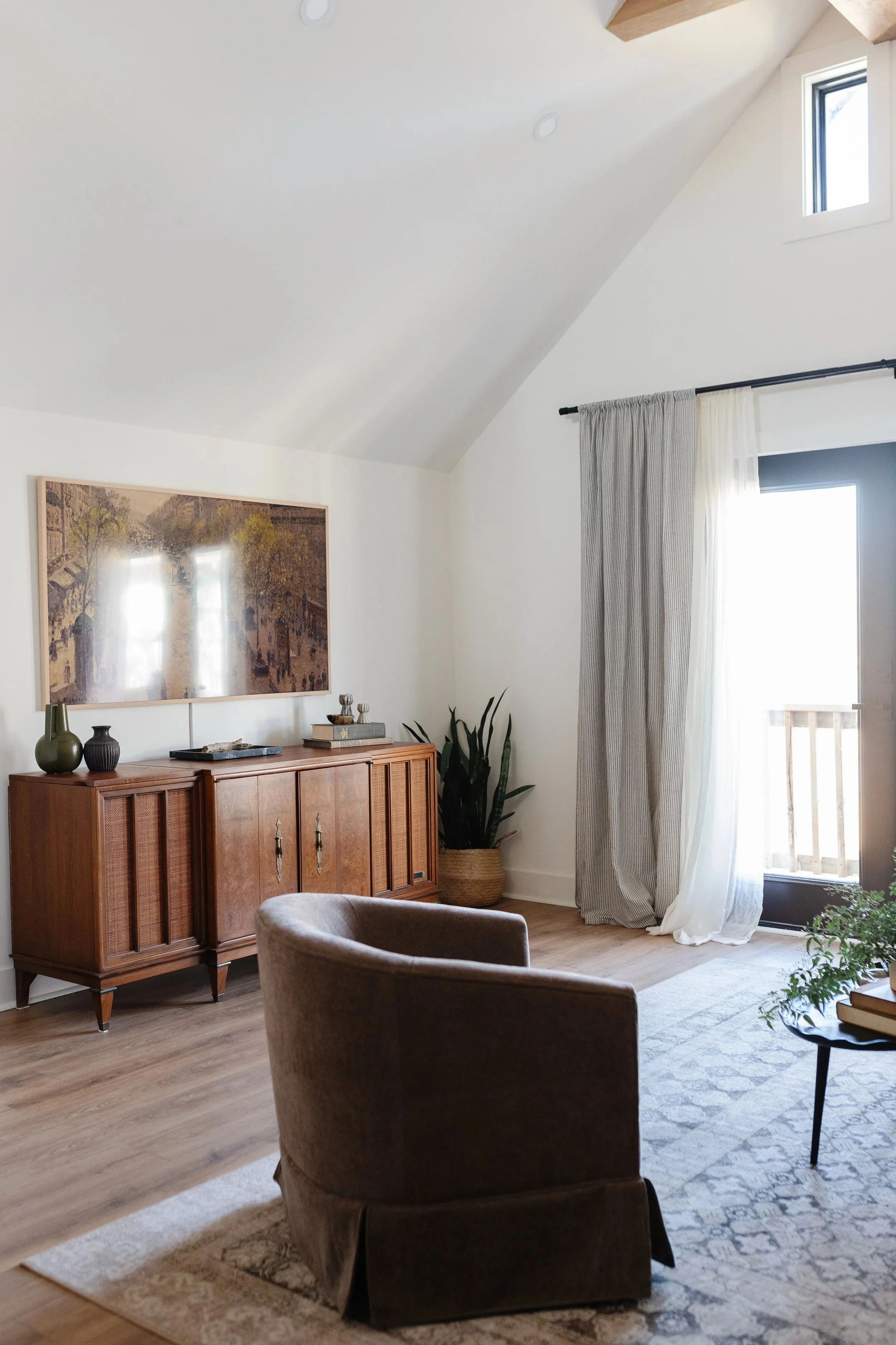 A cozy living room with a wooden sideboard, a potted plant, a rug, and a beige armchair. A large window with sheer and opaque curtains allows natural light to brighten the space.