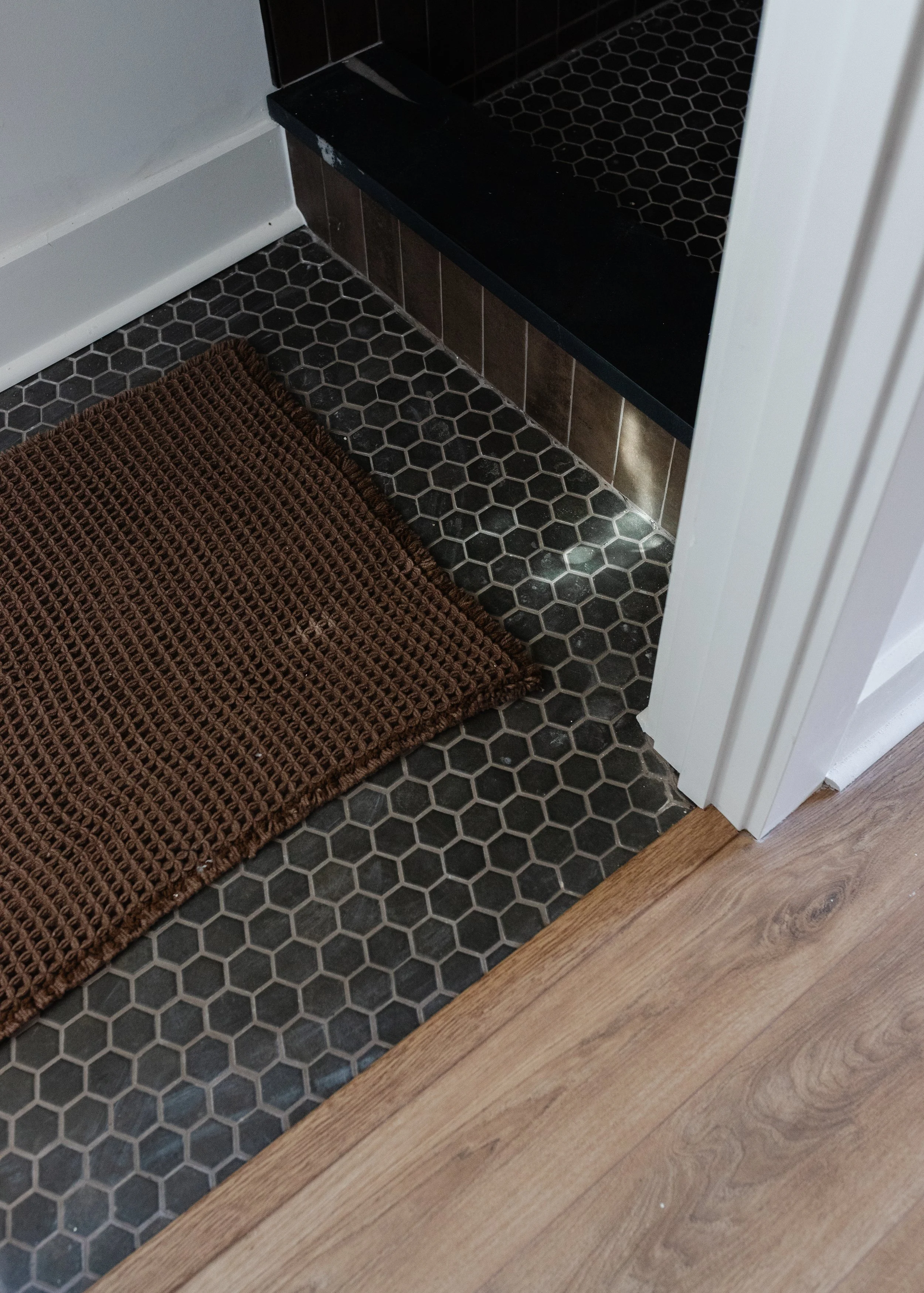 Close-up of a tiled floor with black hexagonal tiles and a brown woven rug near the corner of a room, with part of a doorway and a baseboard.