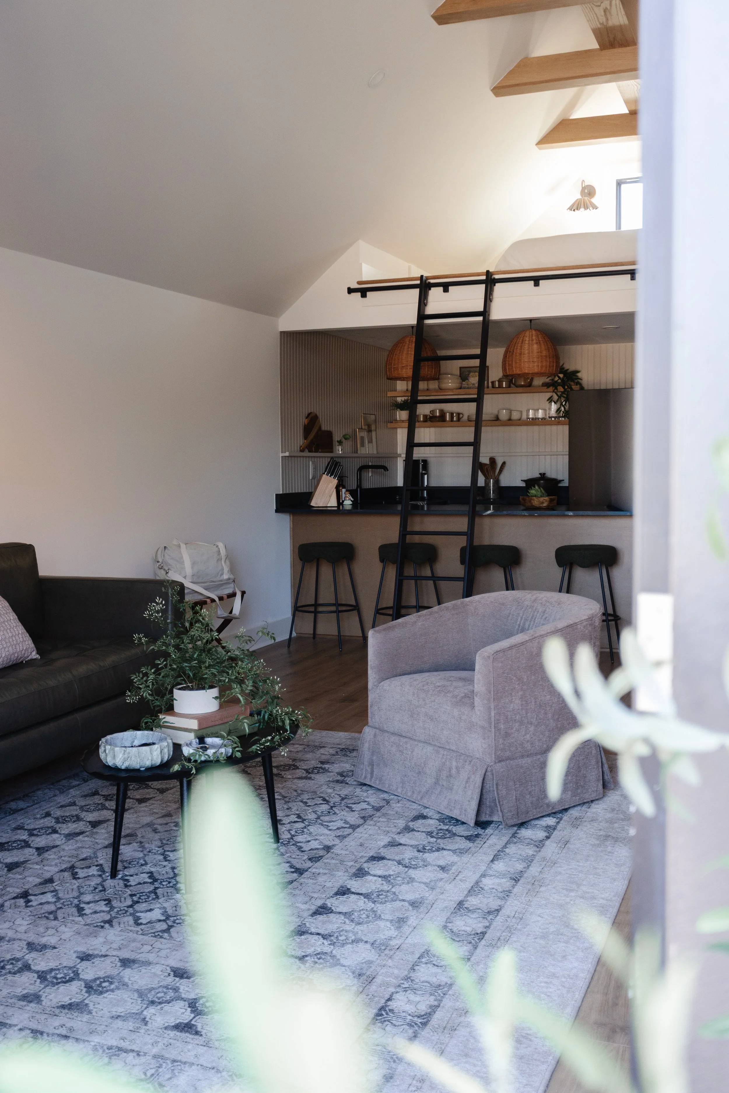 Living room with a black leather sofa, a beige armchair, a black coffee table with plants and books, and a patterned rug. In the background, there is a kitchen with a bar counter, black barstools, open shelving, and hanging woven lamps. A ladder lead