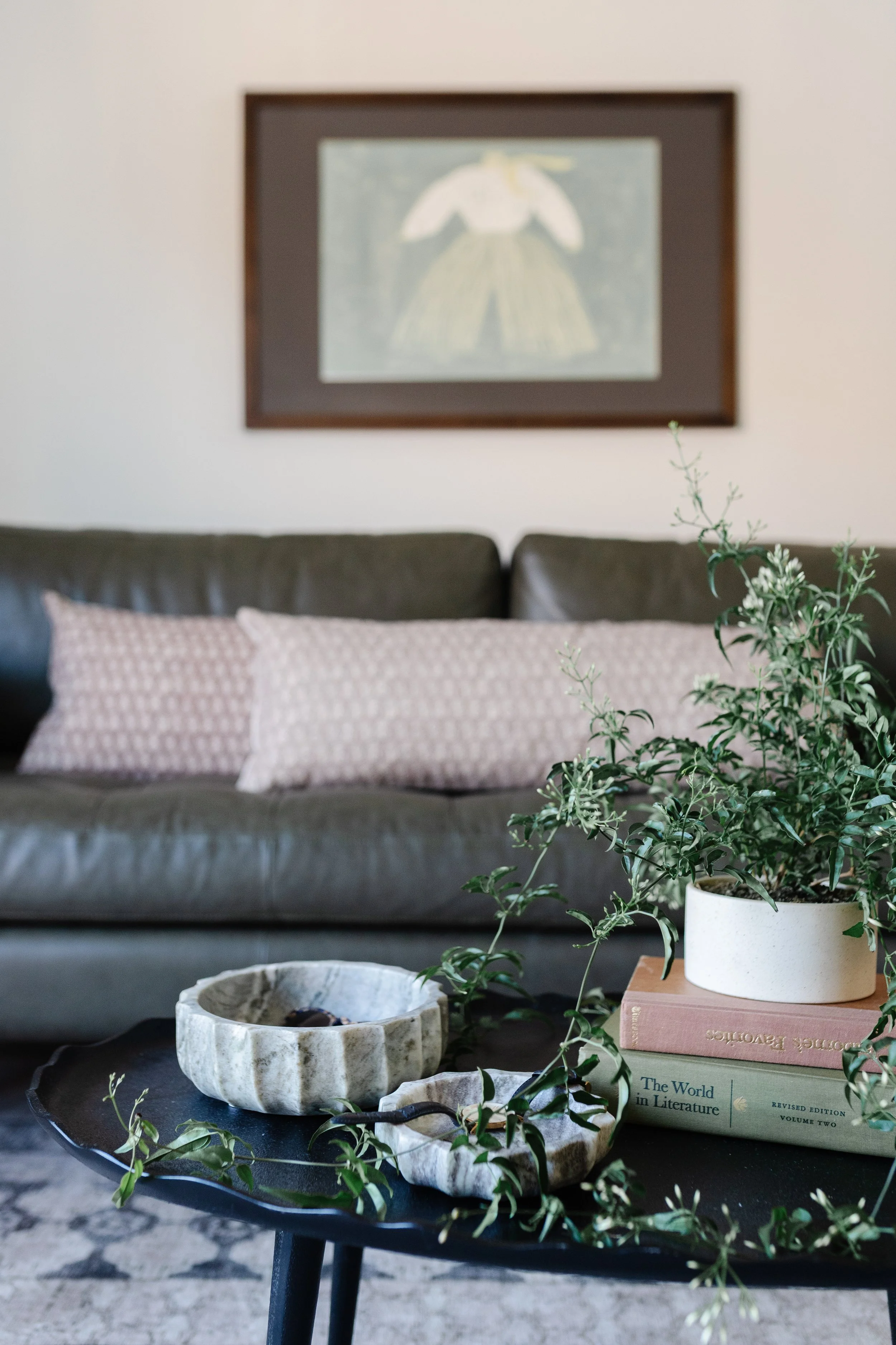 A living room with a dark leather couch, patterned pink pillows, a small black coffee table with marble decor bowls, a potted green plant on a stack of books, and a framed picture of an angel on the wall.