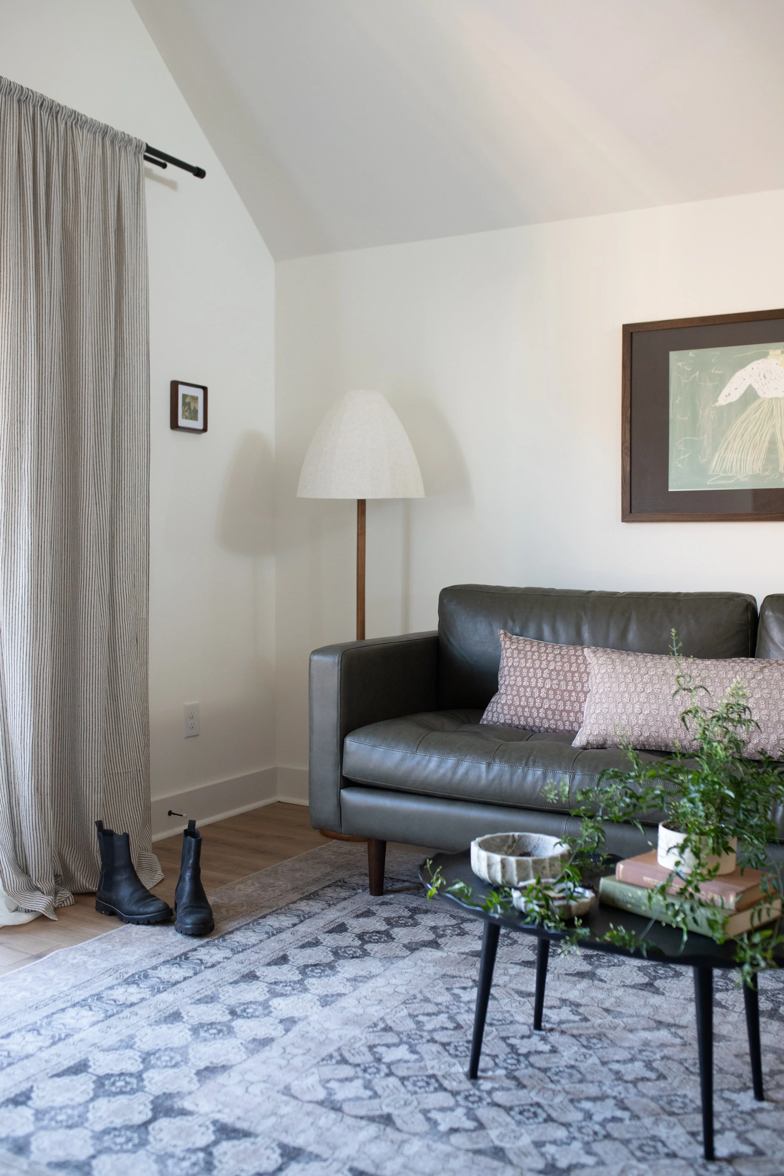 Cozy living room with a green leather sofa, a side table with plants and books, a patterned rug, a floor lamp, framed artwork on the wall, a window with striped curtains, and a pair of black boots on the floor.