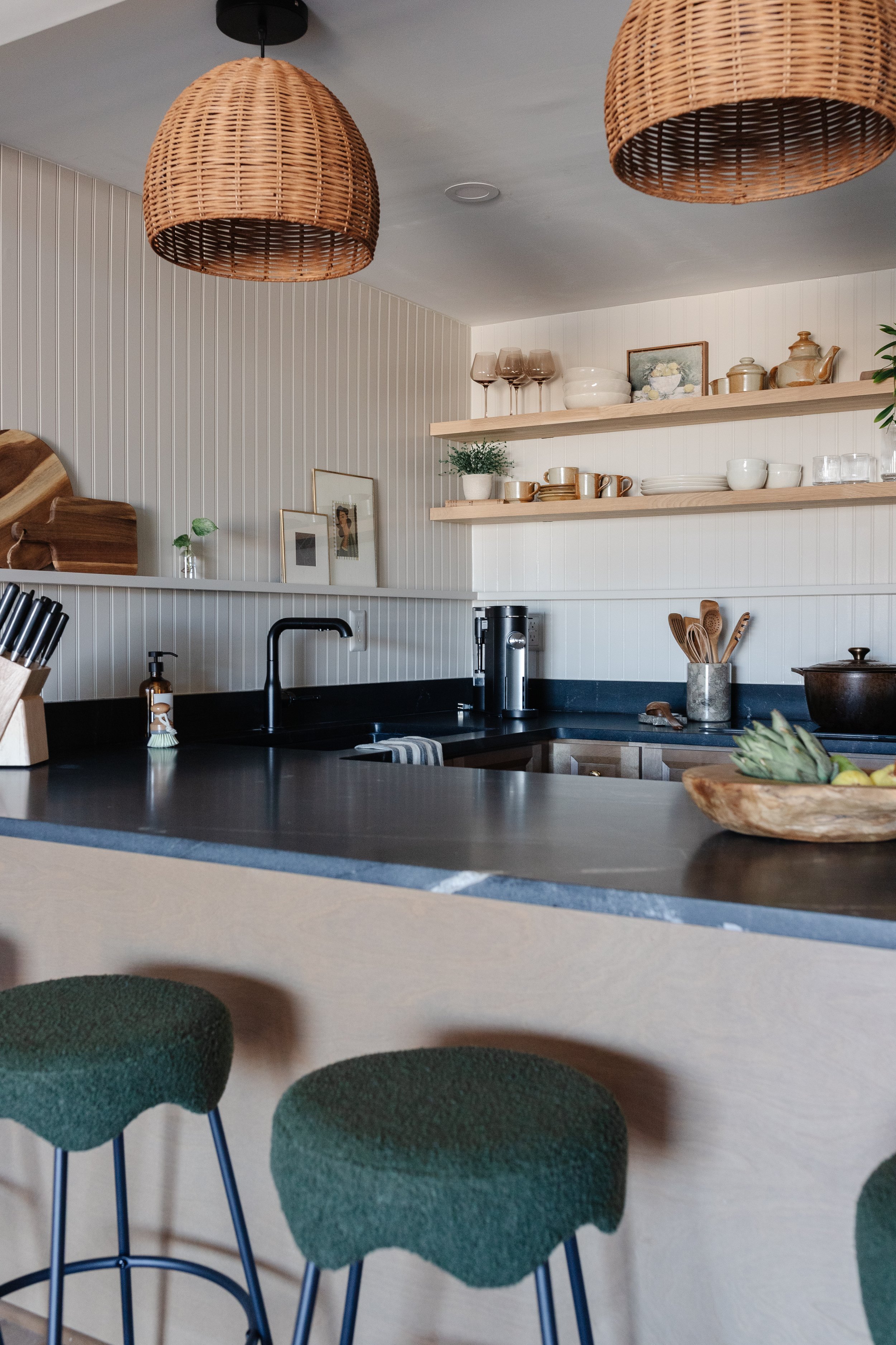 A modern kitchen with black countertops, open wooden shelves with dishes and decor, two large woven pendant lights, green stools at the counter, and a bowl of fresh fruit on the counter.