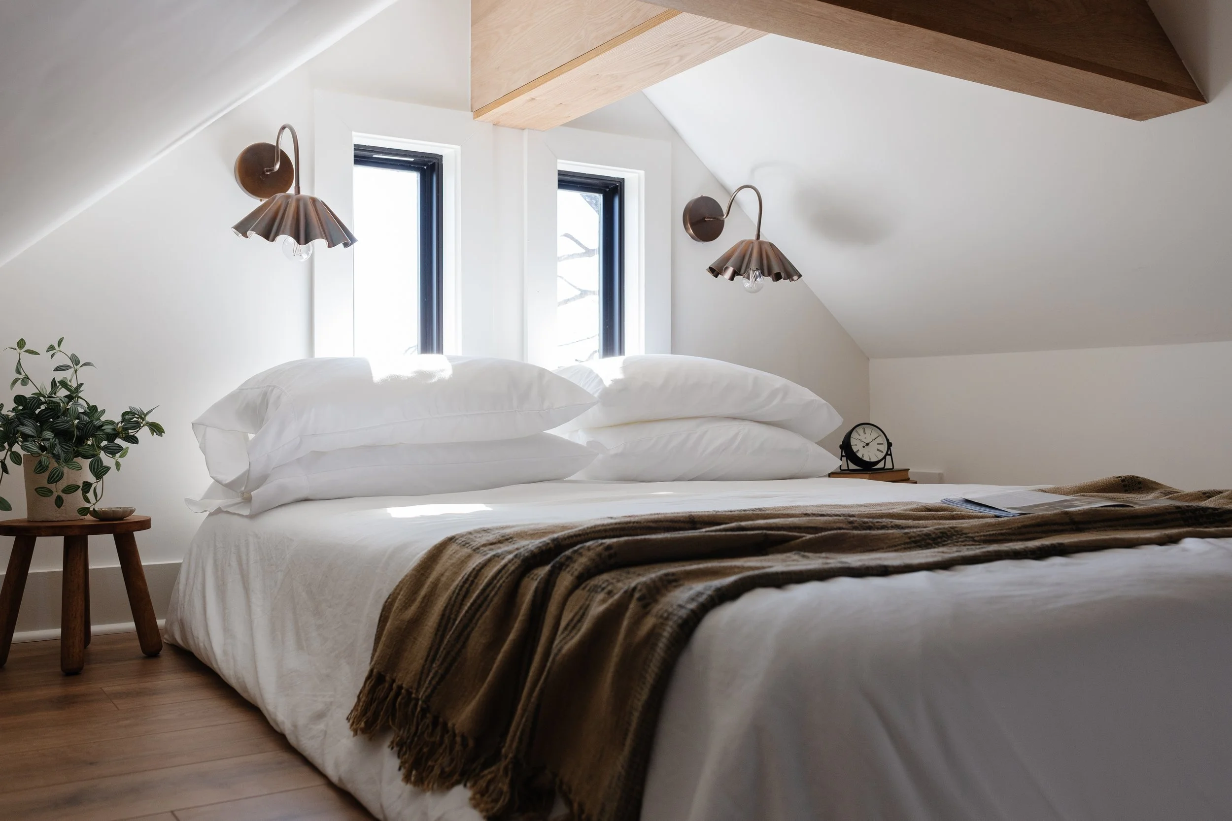 A cozy bedroom with a bed featuring white bedding and multiple pillows, flanked by wall-mounted lamps, two narrow windows, a potted plant on a small wooden side table, and a clock on a nightstand.
