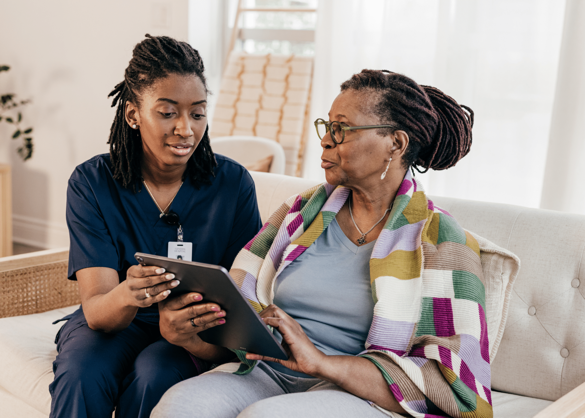 A healthcare worker sitting on a sofa with an elderly woman, showing her information on a tablet in a bright, cozy room.