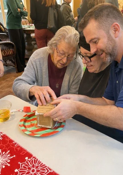 Three people, an elderly woman and two adults, are gathered around a table decorating a gingerbread house for a holiday celebration.