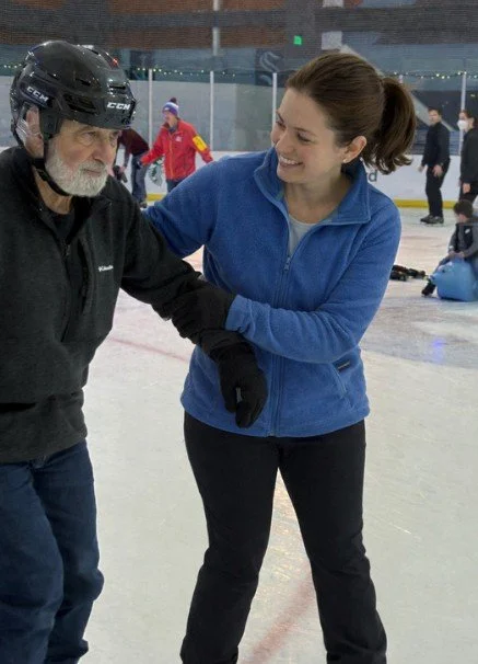 A young woman in a blue jacket helping an older man with a hockey helmet on inside an ice skating rink.