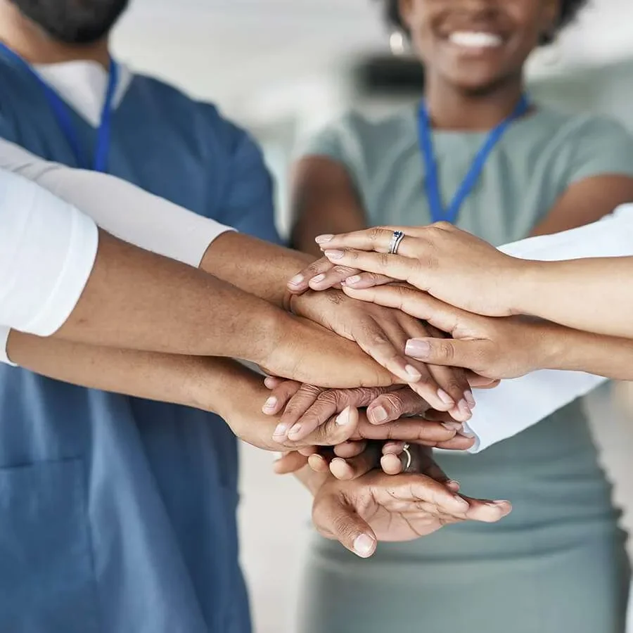 A group of diverse people with different skin tones placing their hands together in unity, smiling in a collaborative environment.