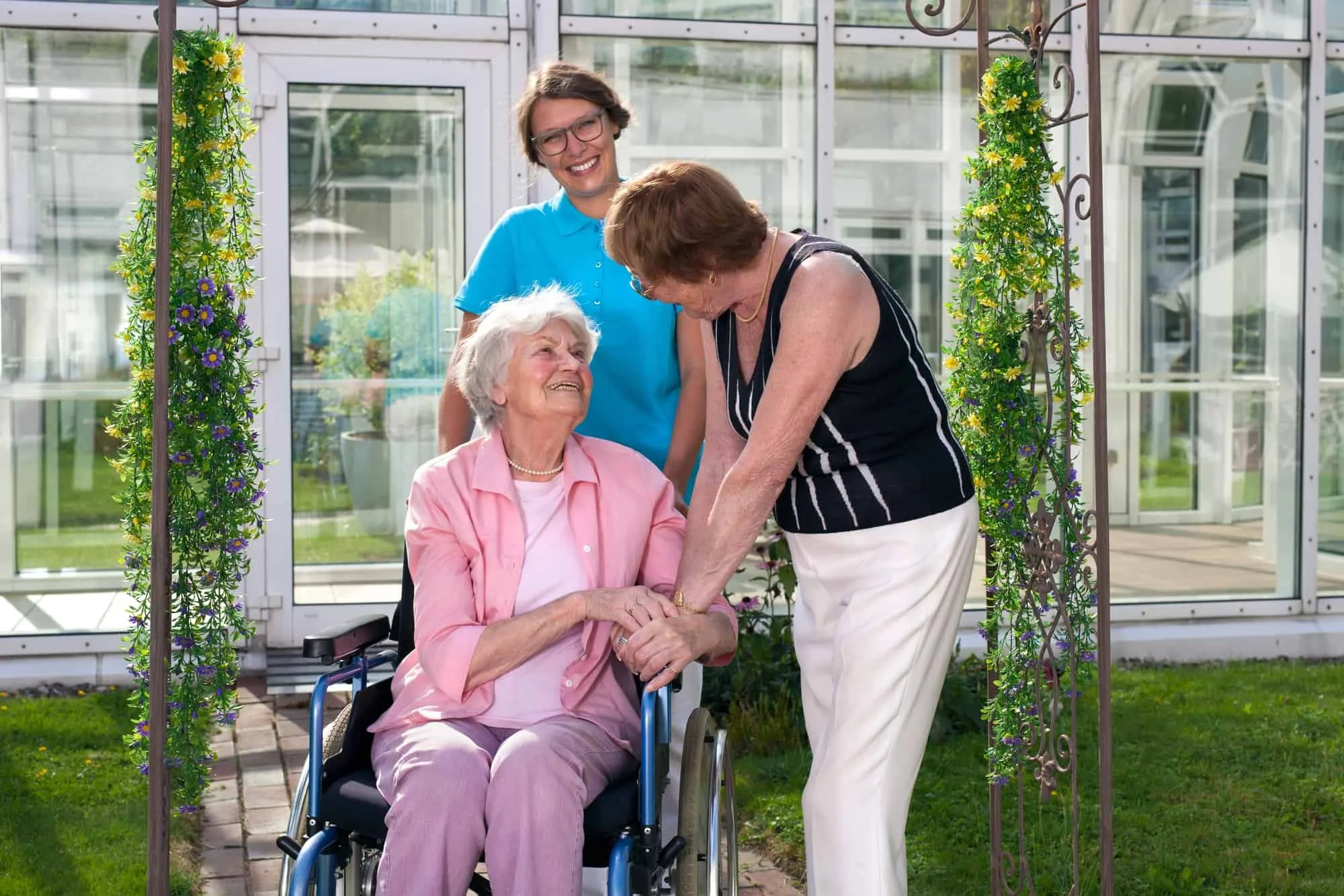 Three women, including an elderly woman in a wheelchair, standing outside a greenhouse with floral decorations, smiling and holding hands.