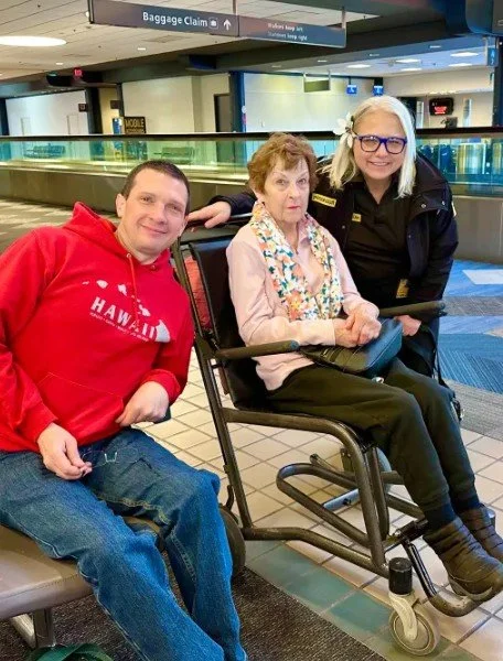 A young man in a red hoodie and an older woman in a wheelchair are at an airport baggage claim area, with a staff member standing beside them.