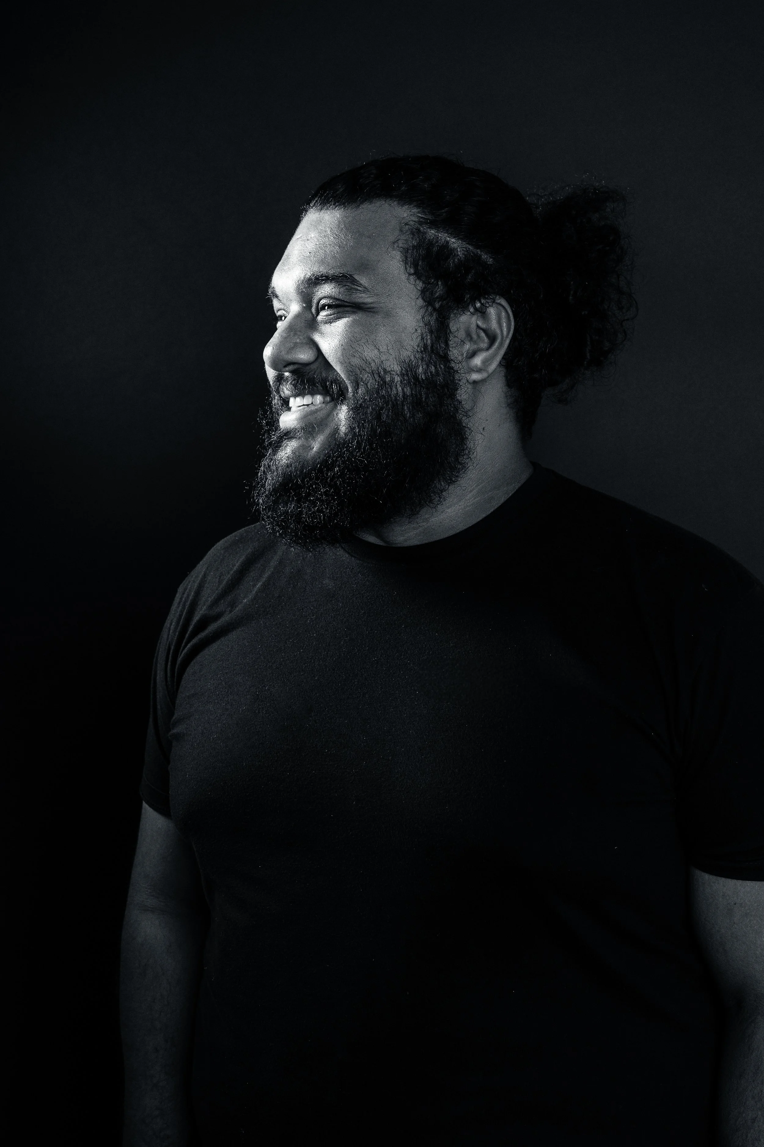A smiling man with a beard and long hair in a bun, wearing a black t-shirt against a dark background. Riverside Portraits